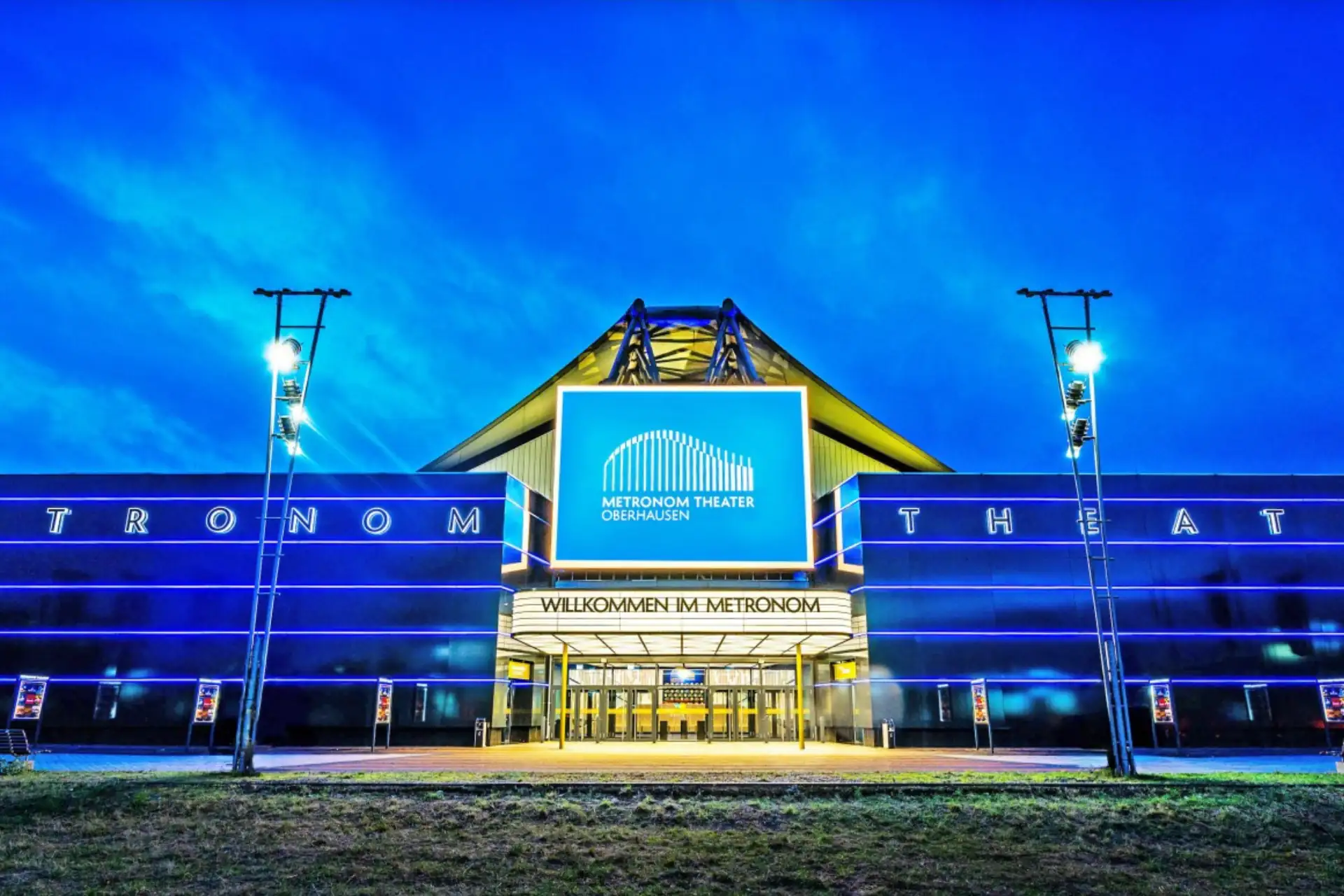 Metronom Theater Oberhausen von außen: Eingang des Metronom Theaters in Oberhausen bei Abenddämmerung mit beleuchtetem Schriftzug 'Metronom Theater' und Willkommensbanner