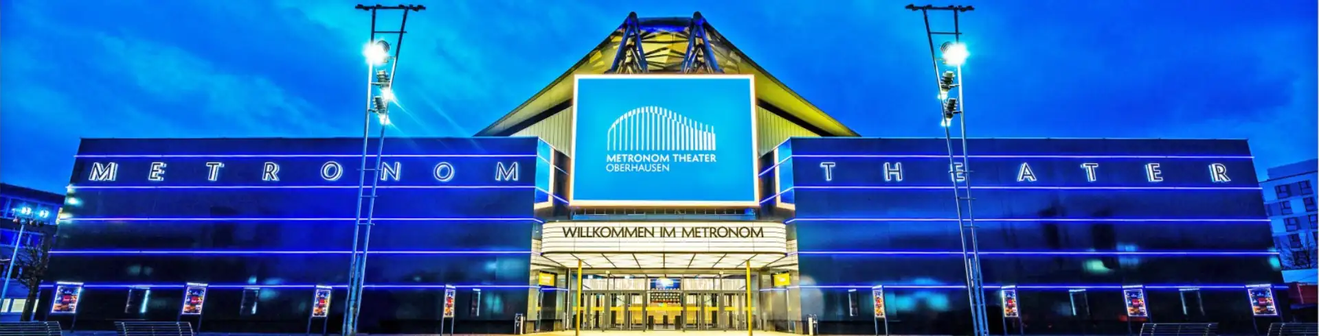 Eingang des Metronom Theaters in Oberhausen bei Abenddämmerung mit beleuchtetem Schriftzug 'Metronom Theater' und Willkommensbanner