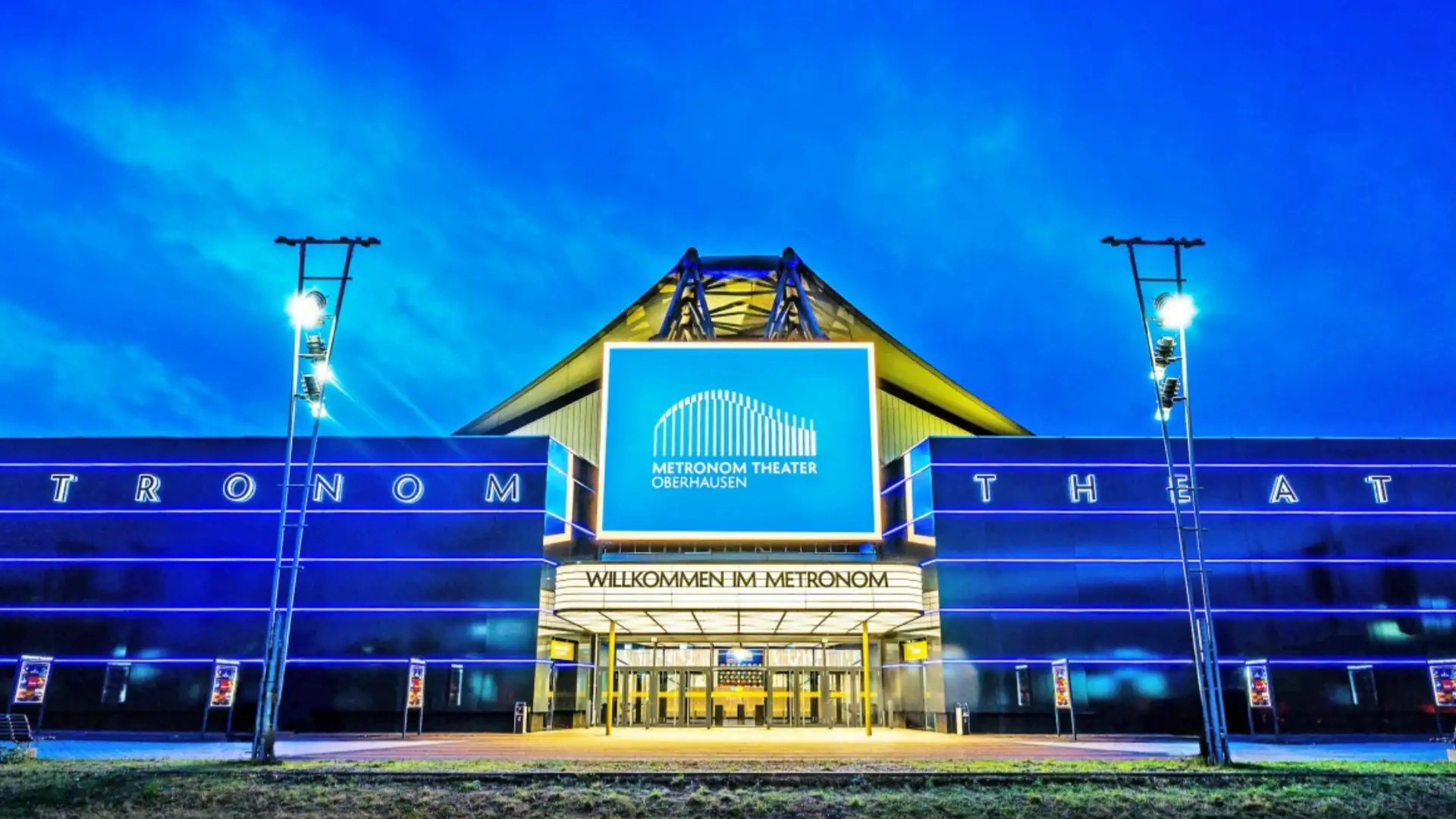 Eingang des Metronom Theaters Oberhausen bei Abenddämmerung mit beleuchtetem Schriftzug und Willkommensschild (im Kachelformat 1920x1080px)