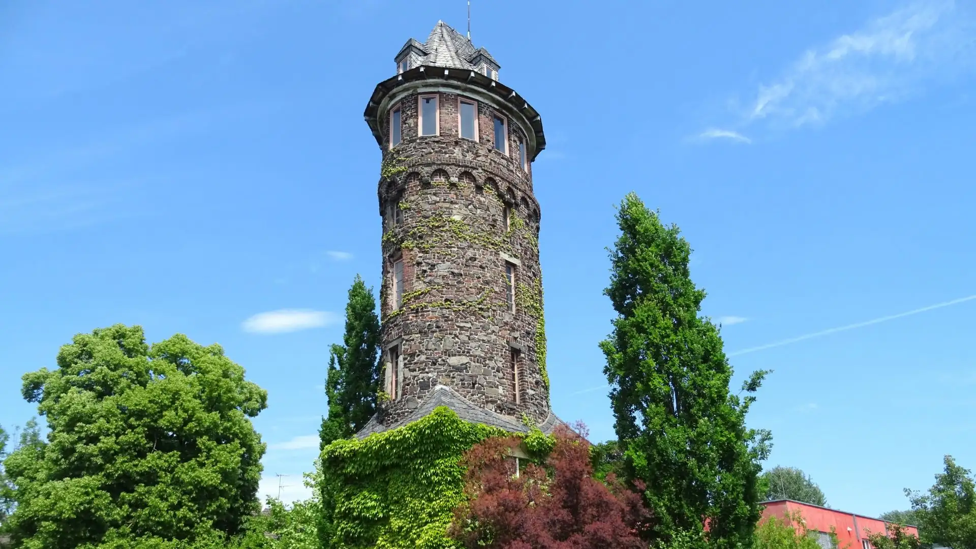 Ein historischer Steinturm umgeben von grünen Bäumen und blauem Himmel.