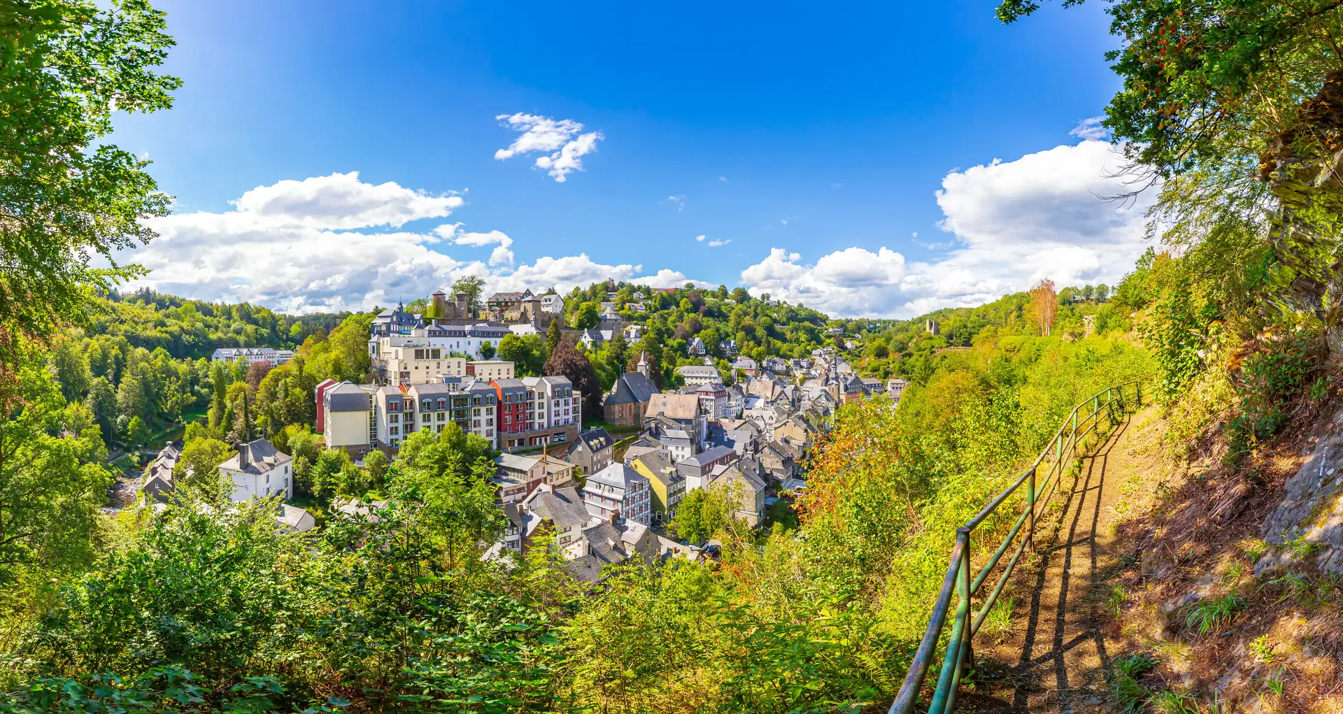 Panoramablick auf Monschau mit historischen Gebäuden und umliegendem Wald unter blauem Himmel