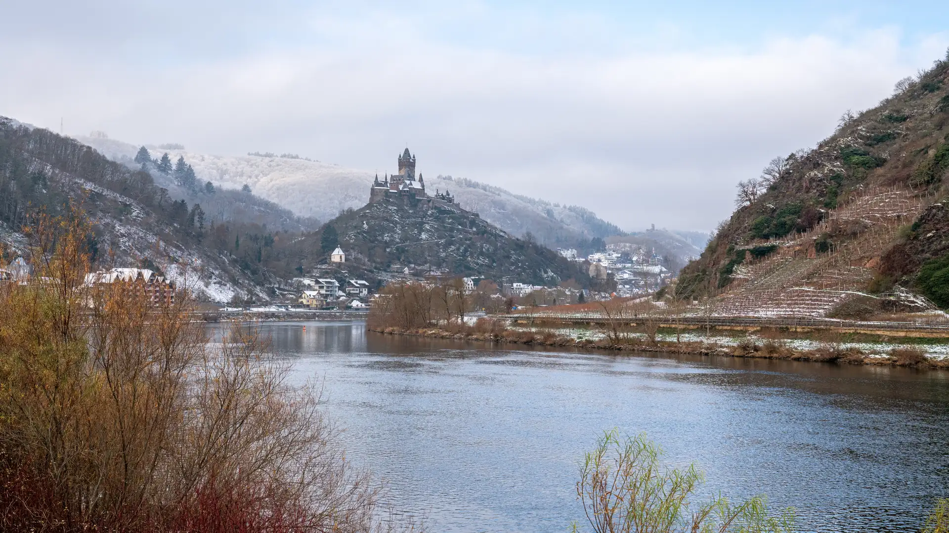 Winterpanorama der Mosel bei Cochem mit der Reichsburg auf einem Hügel und schneebedeckten Weinbergen