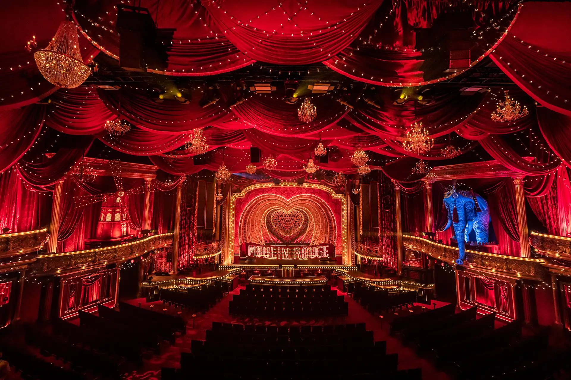 Moulin Rouge! Das Musical in Köln: opulente Bühne mit stimmungsvoller Beleuchtung, rot samtenen Vorhängen, Kronleuchtern sowie blauem Elefanten (rechts) und Windmühle (links)