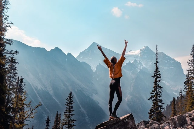 Das Bild zeigt eine Frau, die auf einem Felsen in den Bergen steht und die Arme in einer triumphalen Geste in den Himmel streckt. Im Hintergrund sind majestätische Berggipfel und eine weite, bewaldete Landschaft zu sehen. Der Himmel ist klar und es herrscht eine ruhige, kraftvolle Atmosphäre. Die Szene vermittelt ein Gefühl von Freiheit, Abenteuer und der Freude an der Natur, während die Person ihre Erreichung eines Ziels oder eine persönliche Bestätigung feiert.