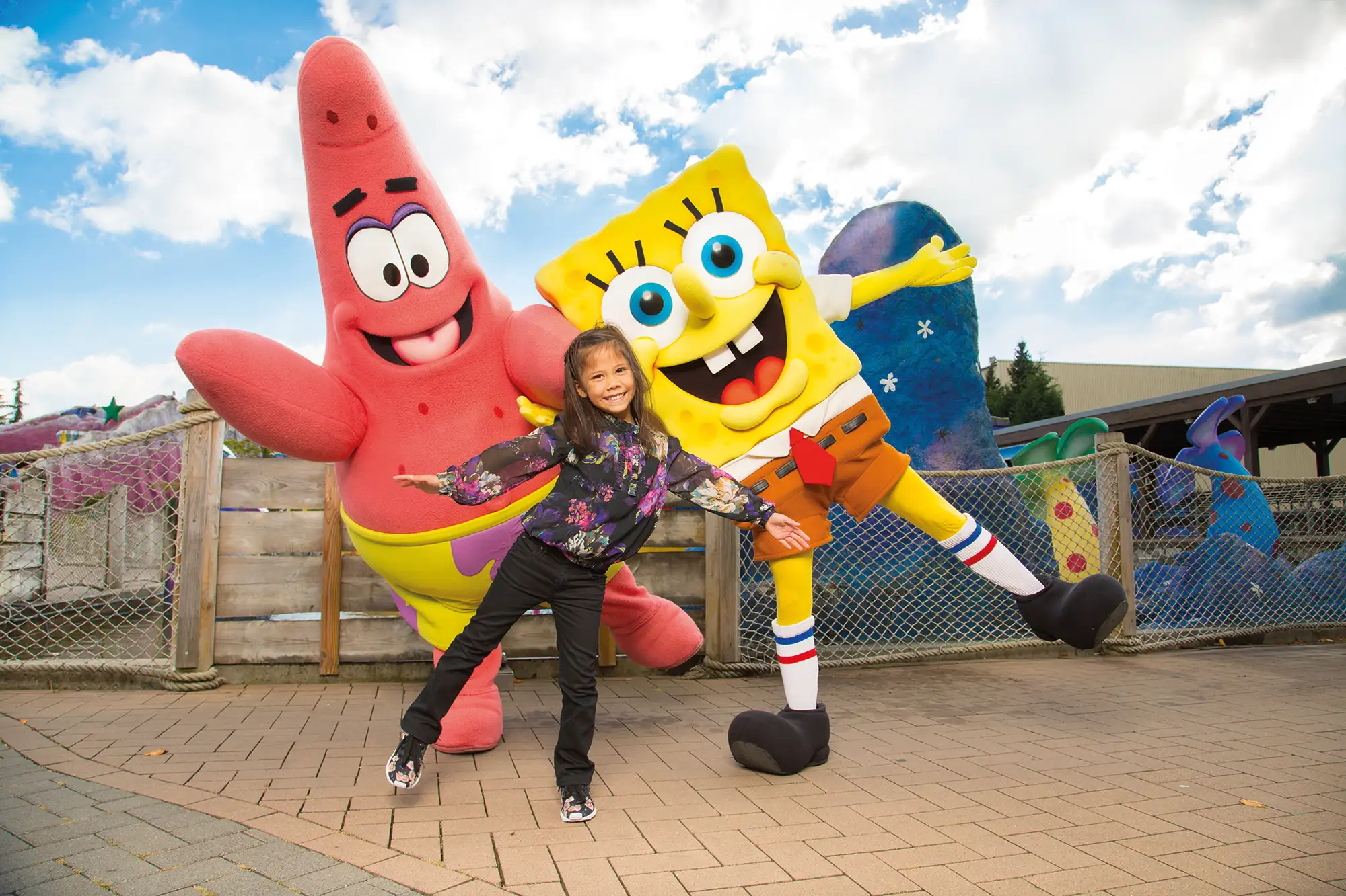 Movie Park mit Übernachtung - SpongeBob und Patrick mit einem Mädchen lachend und auf einem Bein vor einer Achterbahn im Movie Park