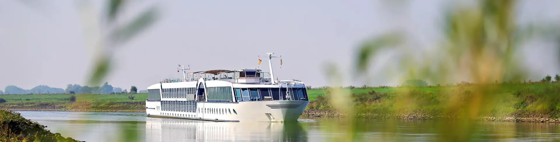 MS Dutch Grace im Bannerformat: Weißes Flusskreuzfahrtschiff fährt auf ruhigem Fluss, umgeben von grünen Uferpflanzen und Wiesen.