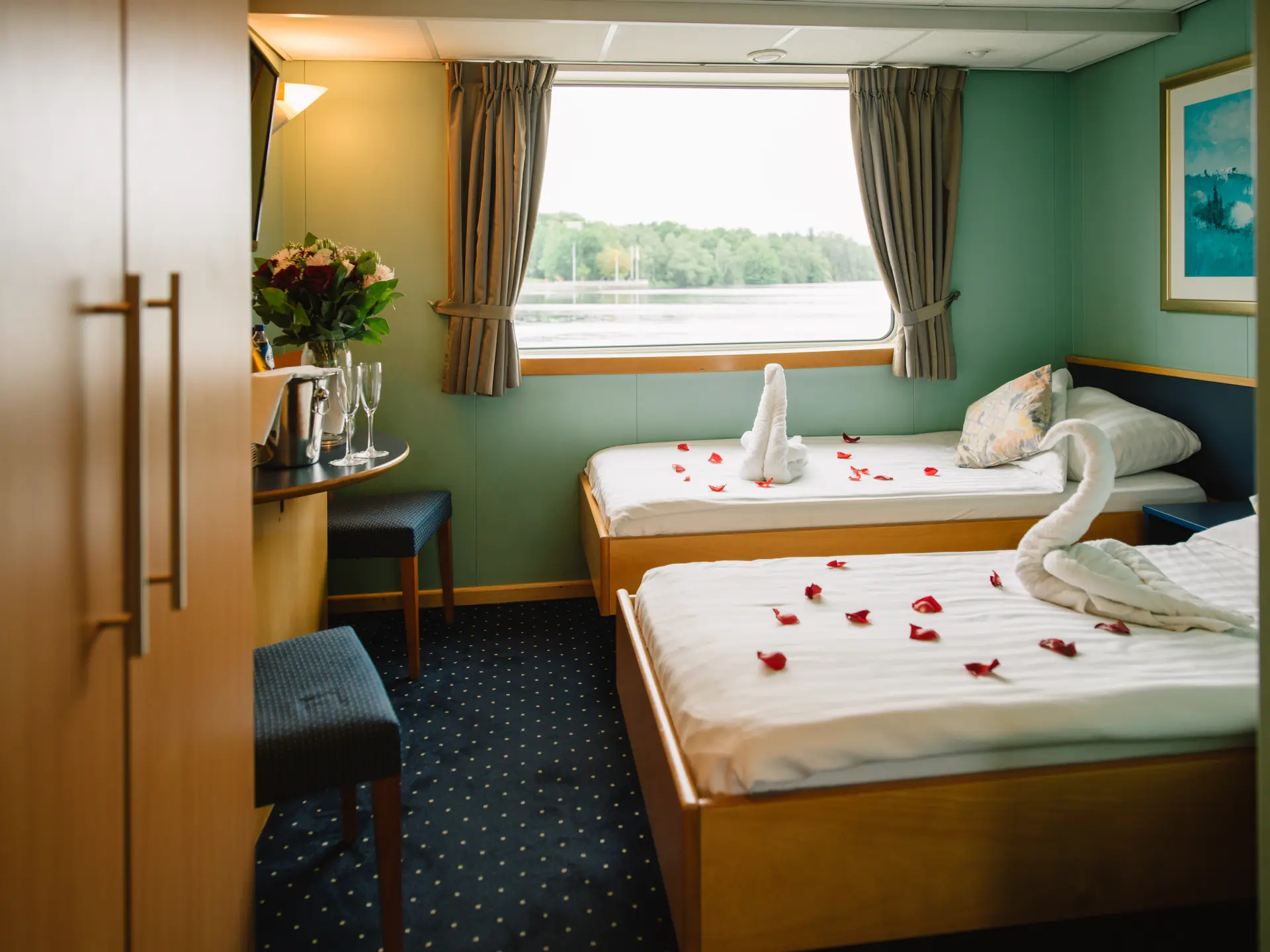 2-Bettkabine auf MS Johannes Brahms: Einzelbetten mit Rosenblättern und Handtuchschwänen dekoriert, Sektgläser auf Schreibtisch und Blick aus dem Kabinenfenster auf die Flusslandschaft