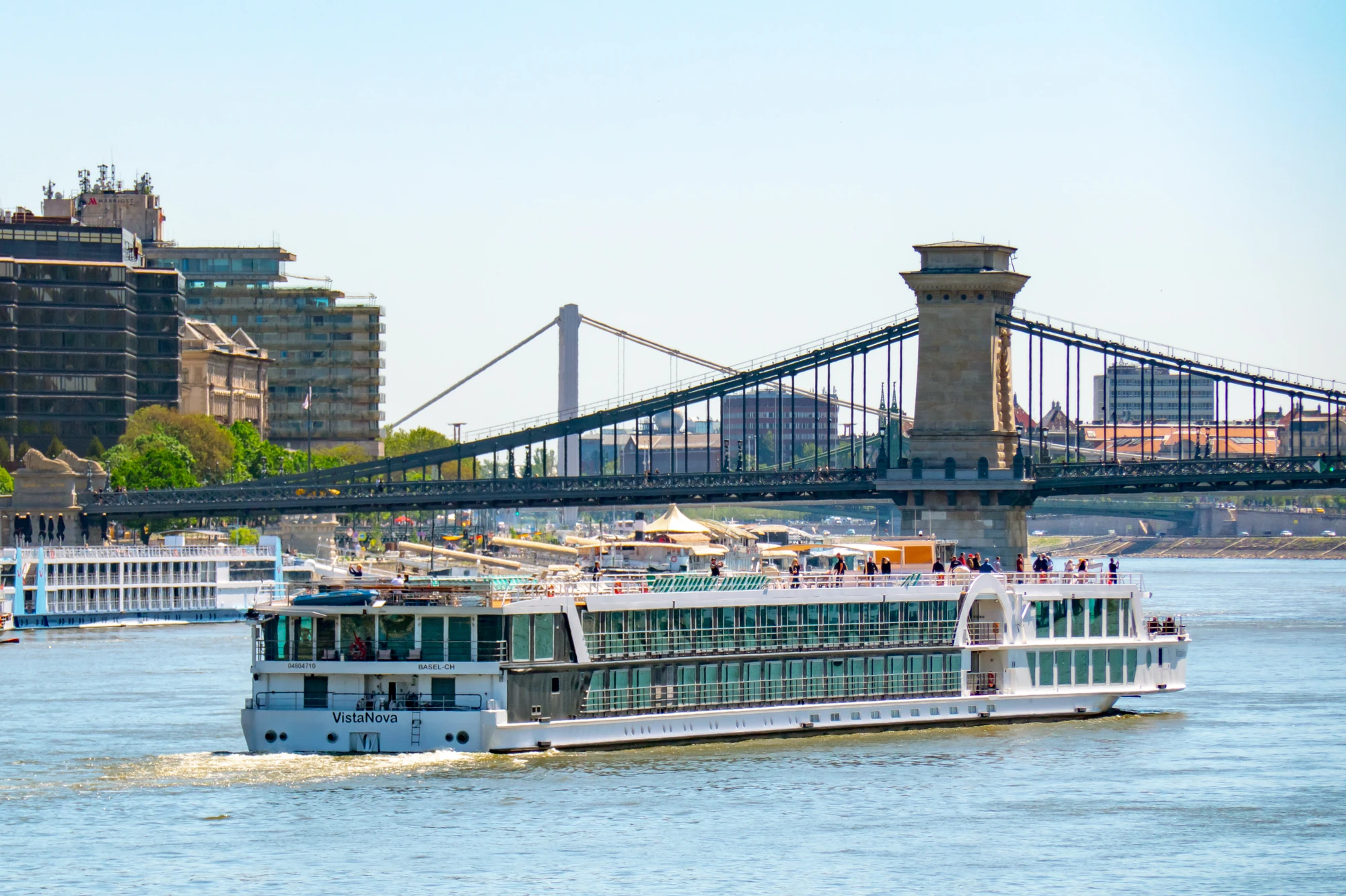 Außenansicht der VistaNova mit mehreren Decks auf einem Fluss, im Hintergrund eine Hängebrücke und Stadtgebäude.