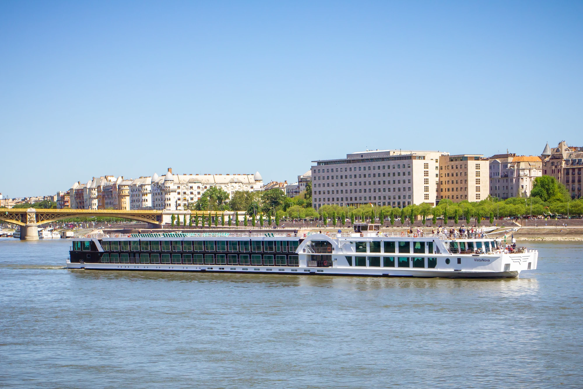 Außenansicht der VistaNova mit mehreren Decks auf ruhigem Fluss vor städtischer Uferkulisse mit Brücke und Gebäuden