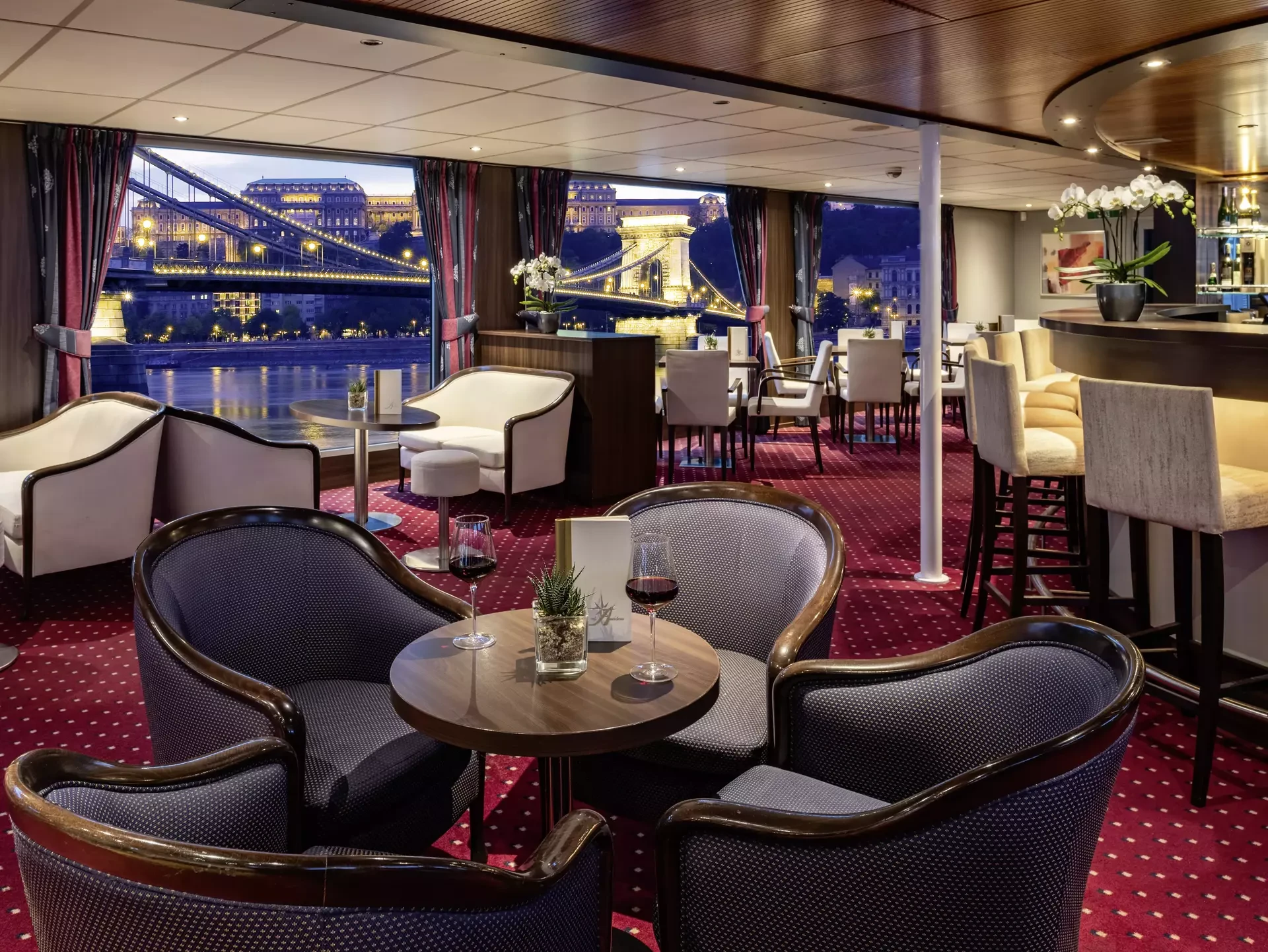 Gemütliche Bar mit runden Tischen, gepolsterten Sesseln und Blick auf die beleuchtete Kettenbrücke bei Nacht auf der VistaNova