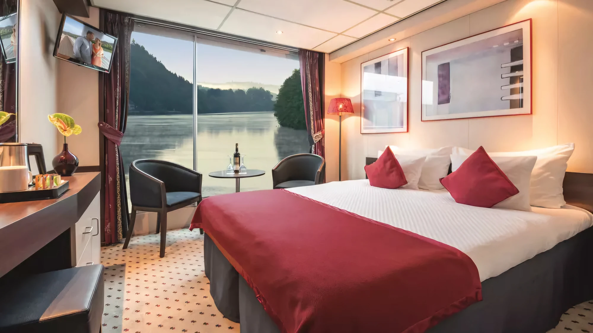 Hotelzimmer mit Doppelbett, rotem Bettläufer und Kissen, zwei Sesseln vor Panoramafenster mit Seeblick, Schreibtisch mit Vase und Lampe