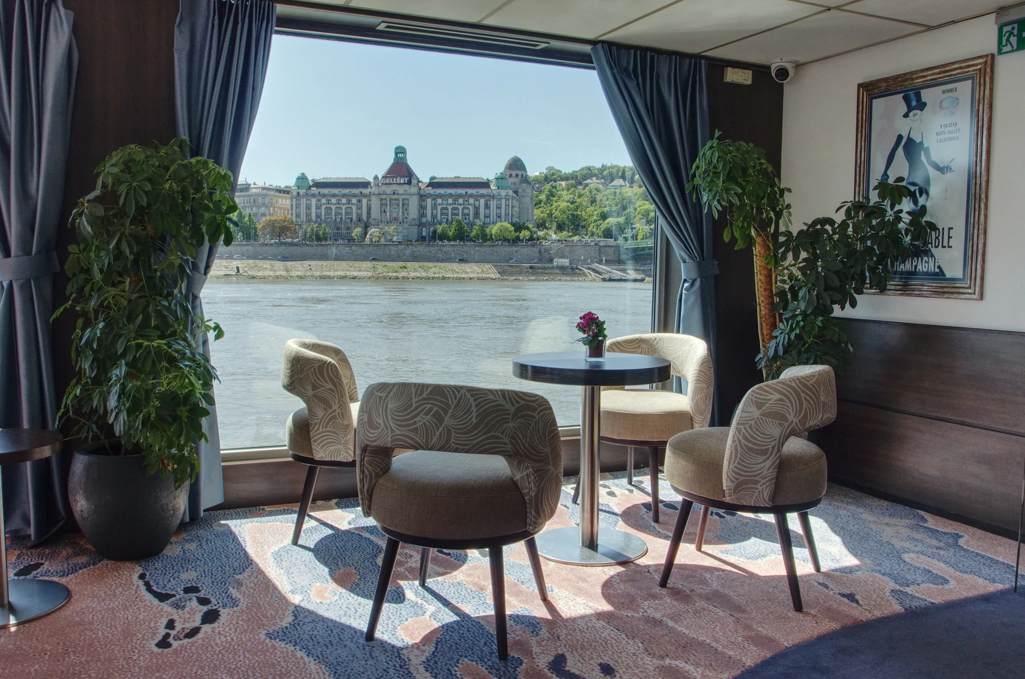 Vier gepolsterte Stühle um einen runden Tisch mit einer kleinen Blume, Blick auf ein Flussufer und ein großes Gebäude durch ein Fenster mit blauen Vorhängen