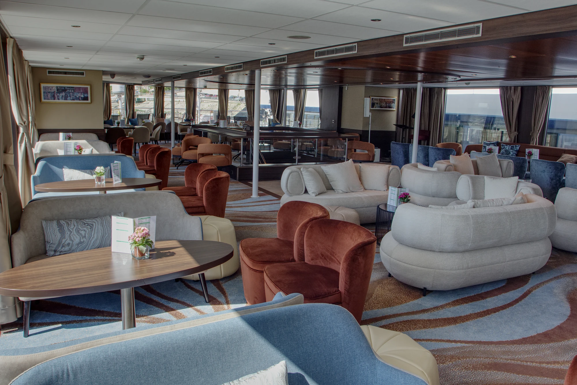 Innenraum eines modernen Lounges mit verschiedenen Sitzgruppen aus Sesseln und Sofas in Blau, Weiß und Braun, Tische mit Blumenvasen und großen Fenstern mit Blick nach draußen