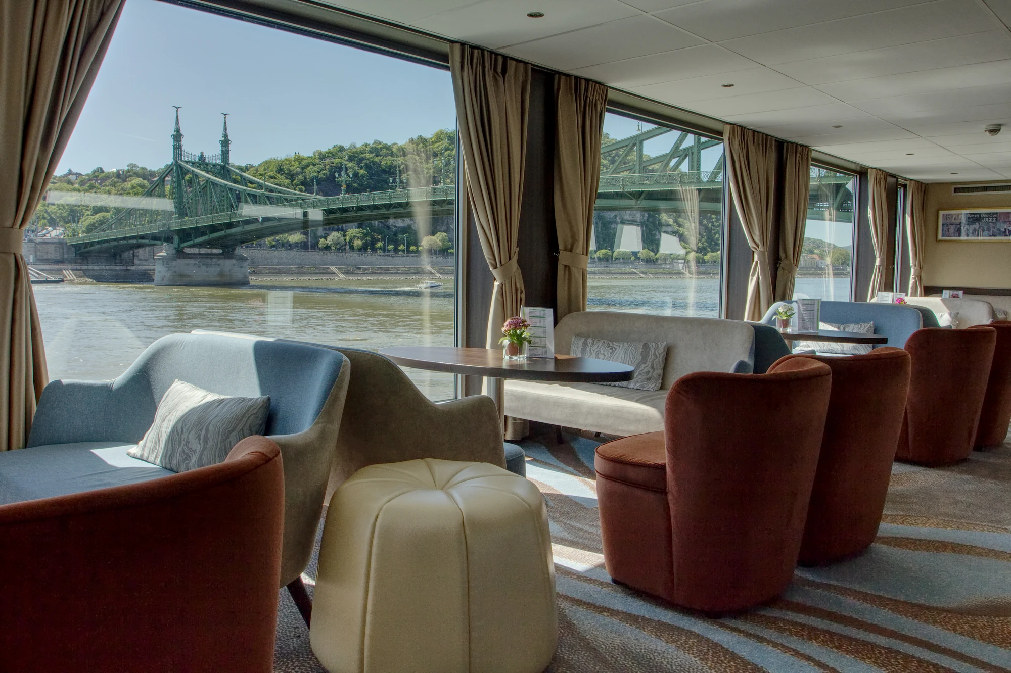 Innenraum mit Sitzgruppen aus Sesseln und Hockern vor großen Fenstern, Blick auf eine grüne Flussuferlandschaft und eine Brücke.