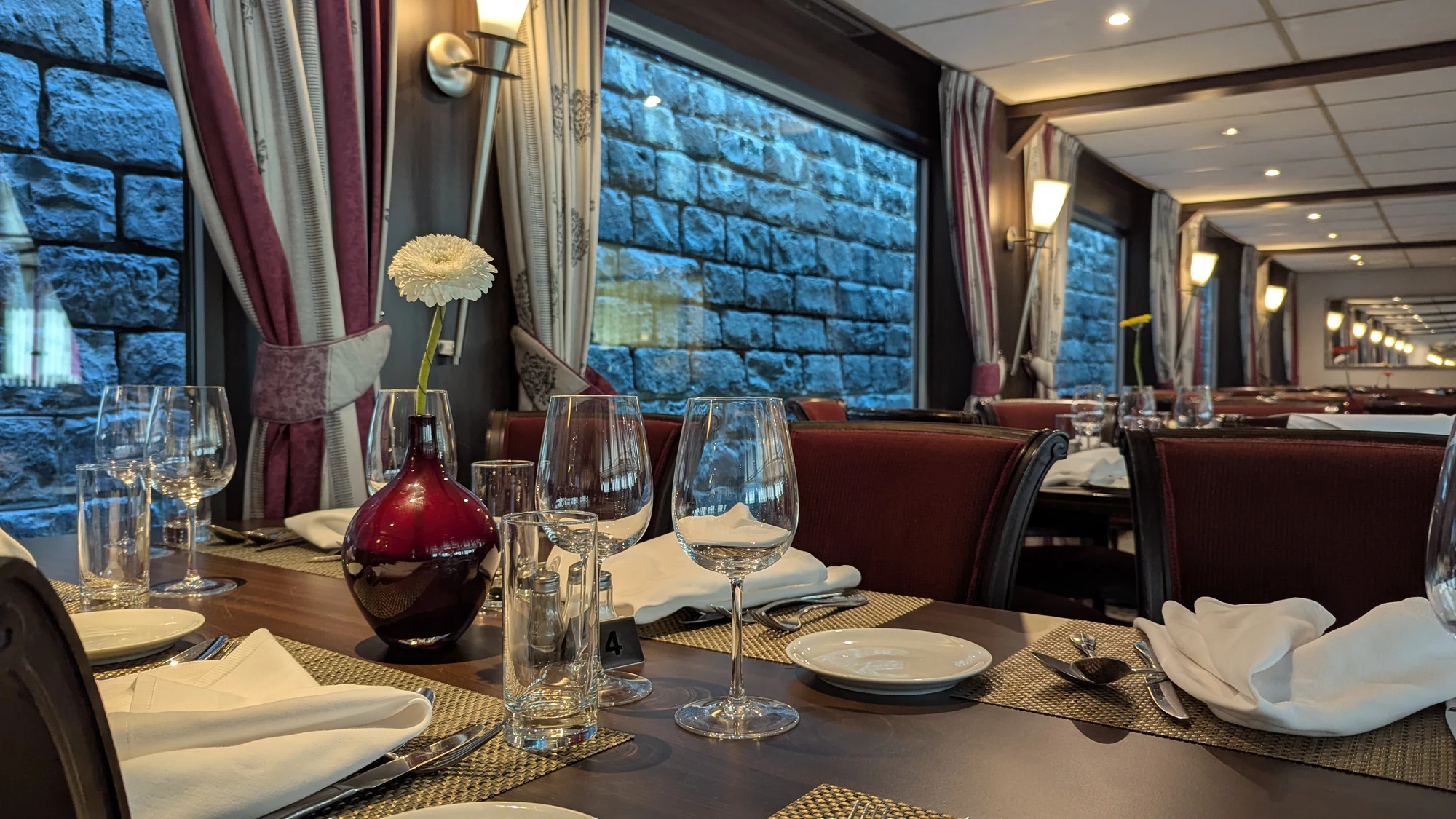 Tisch in einem Restaurant mit rotbrauner Vase, weißer Blume, Weingläsern, Tellern und Besteck vor einer Wand mit blauem Steinmuster auf der VistaNova