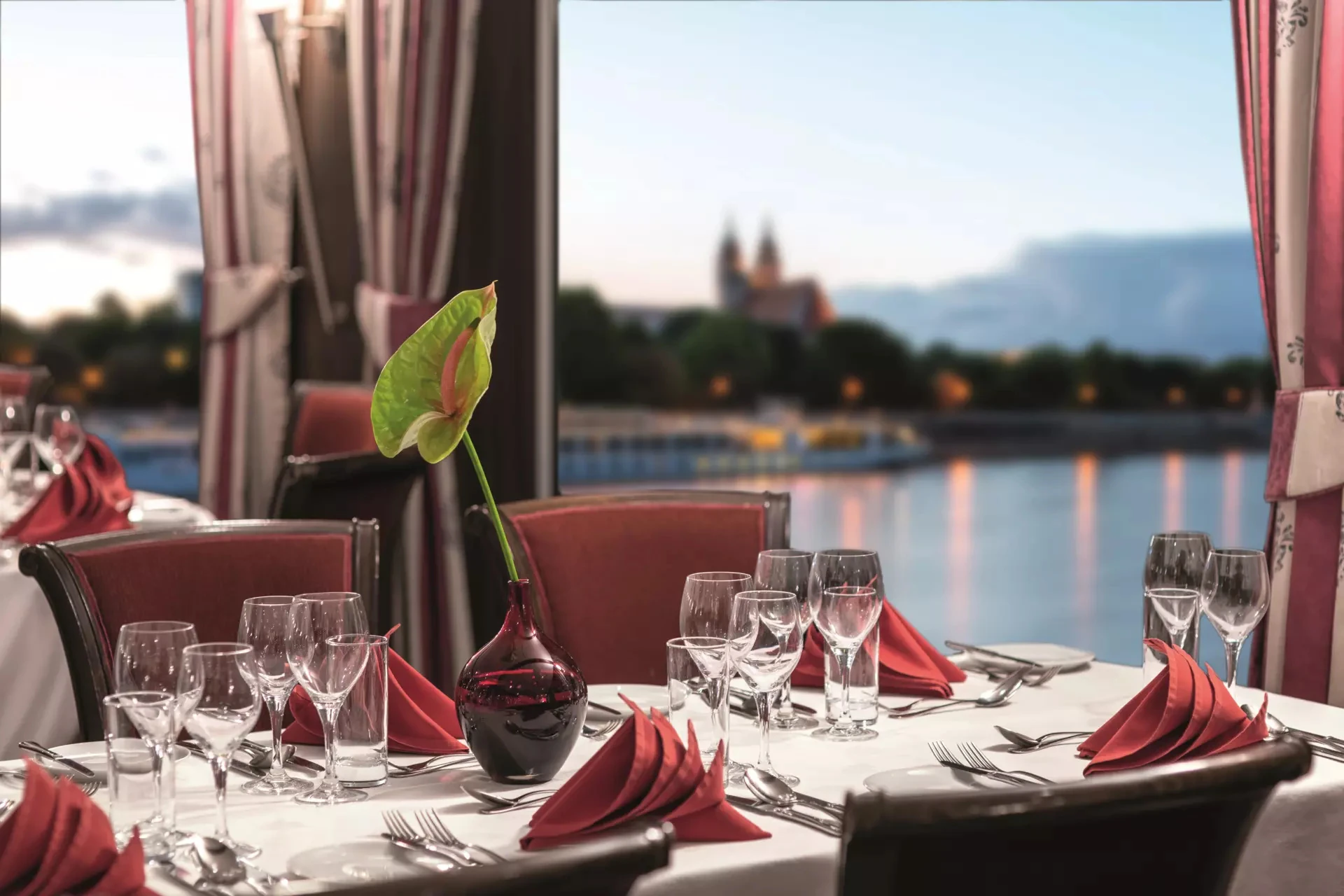 Tisch im Restaurant mit weißer Tischdecke, gefalteten roten Servietten, mehreren Weingläsern und einer grünen Pflanze in einer Vase, im Hintergrund unscharfe Flusslandschaft mit Gebäuden auf der VistaNova