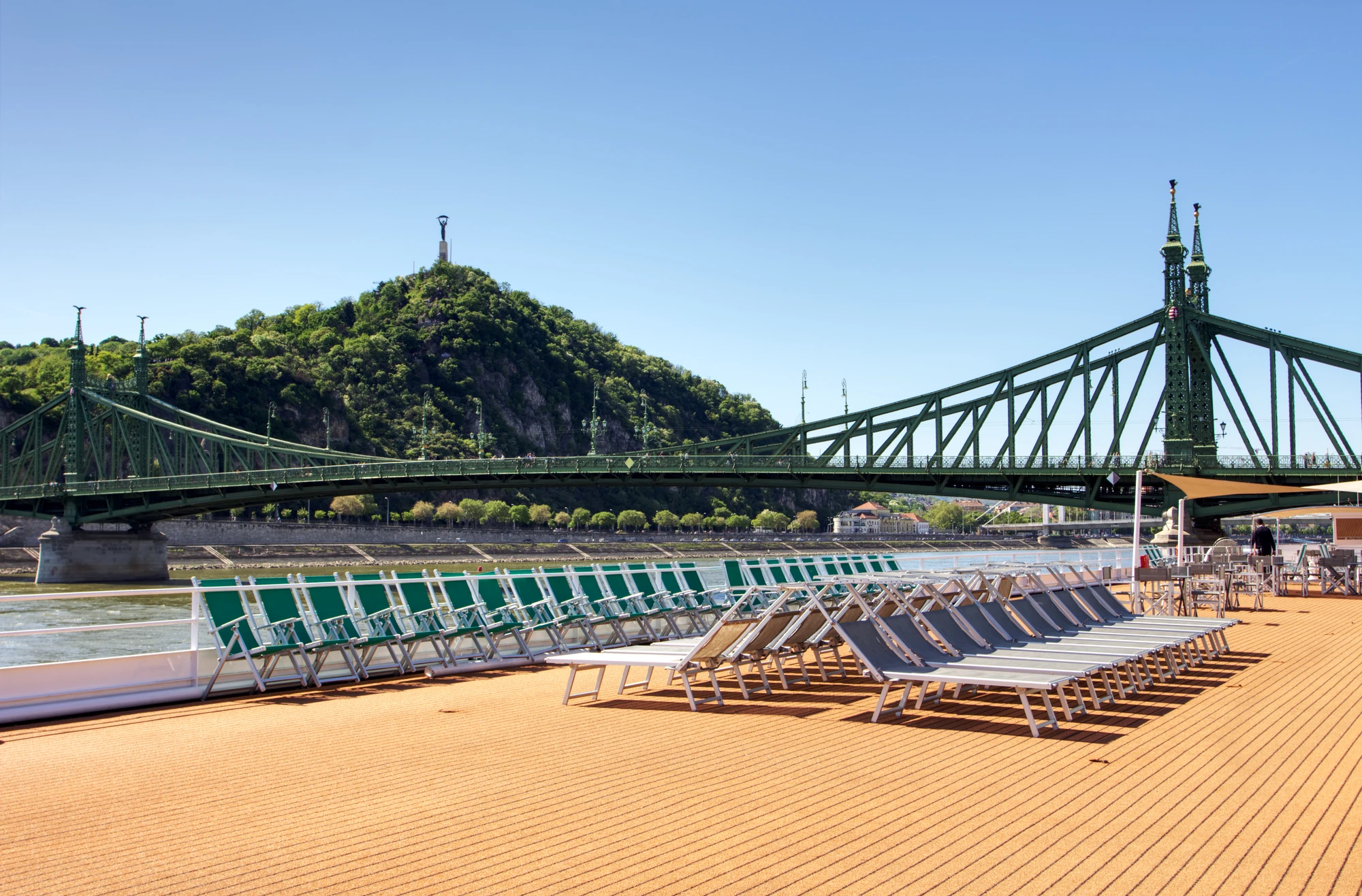 Grüne und weiße Liegestühle auf Holzdeck am Flussufer, im Hintergrund grüner Hügel und grüne Stahlbrücke unter blauem Himmel auf der VistaNova