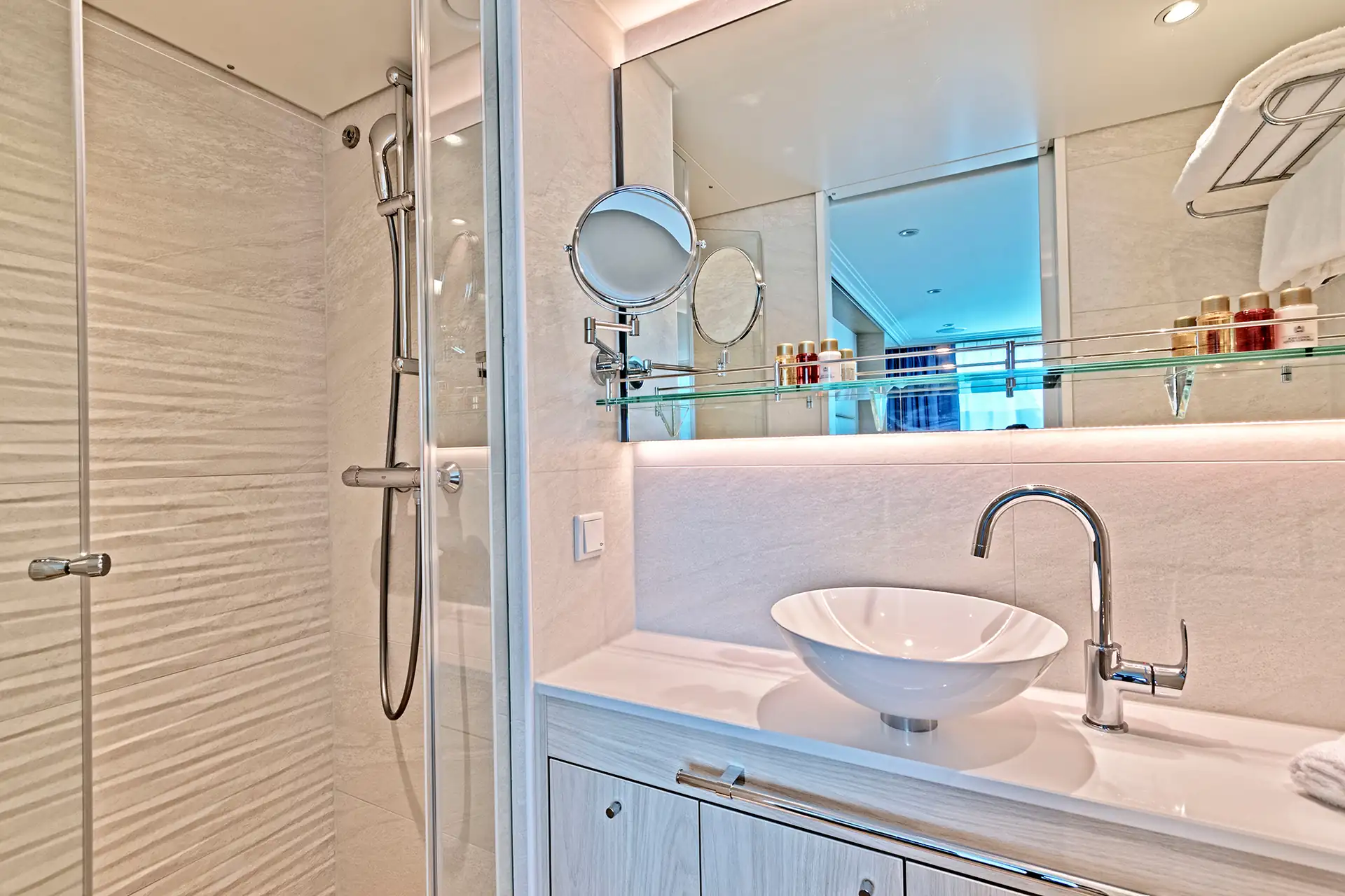 MS VistaSTAR: Weißes Badezimmer mit rundem Waschbecken, verchromtem Wasserhahn, beleuchtetem Spiegel und Duschkabine mit Glaswand und Handbrause.