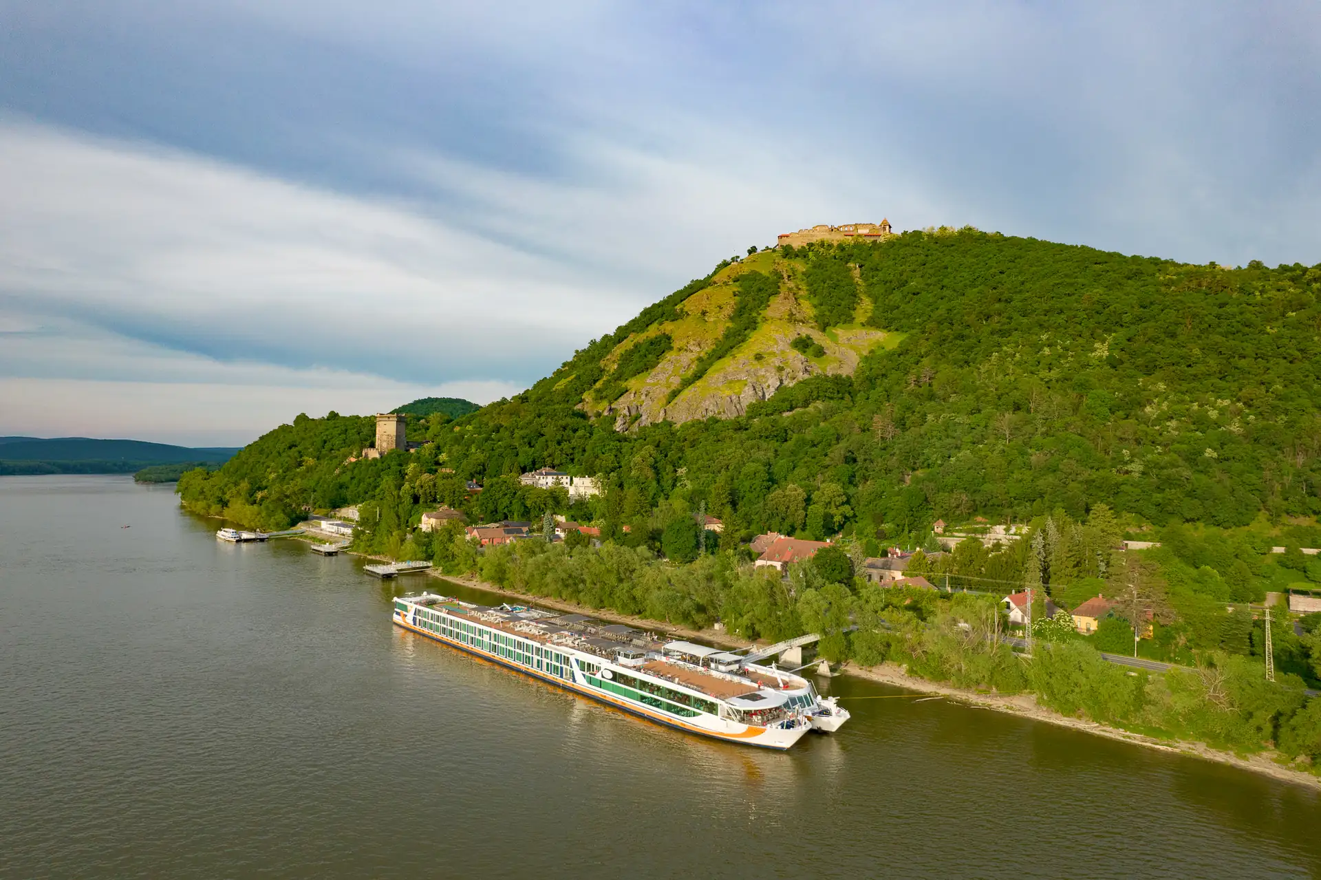 MS VistaSTAR am Ufer der Donau mit bewaldeten Hügel, Ruine und Gebäude auf dem Gipfel unter bewölktem Himmel.