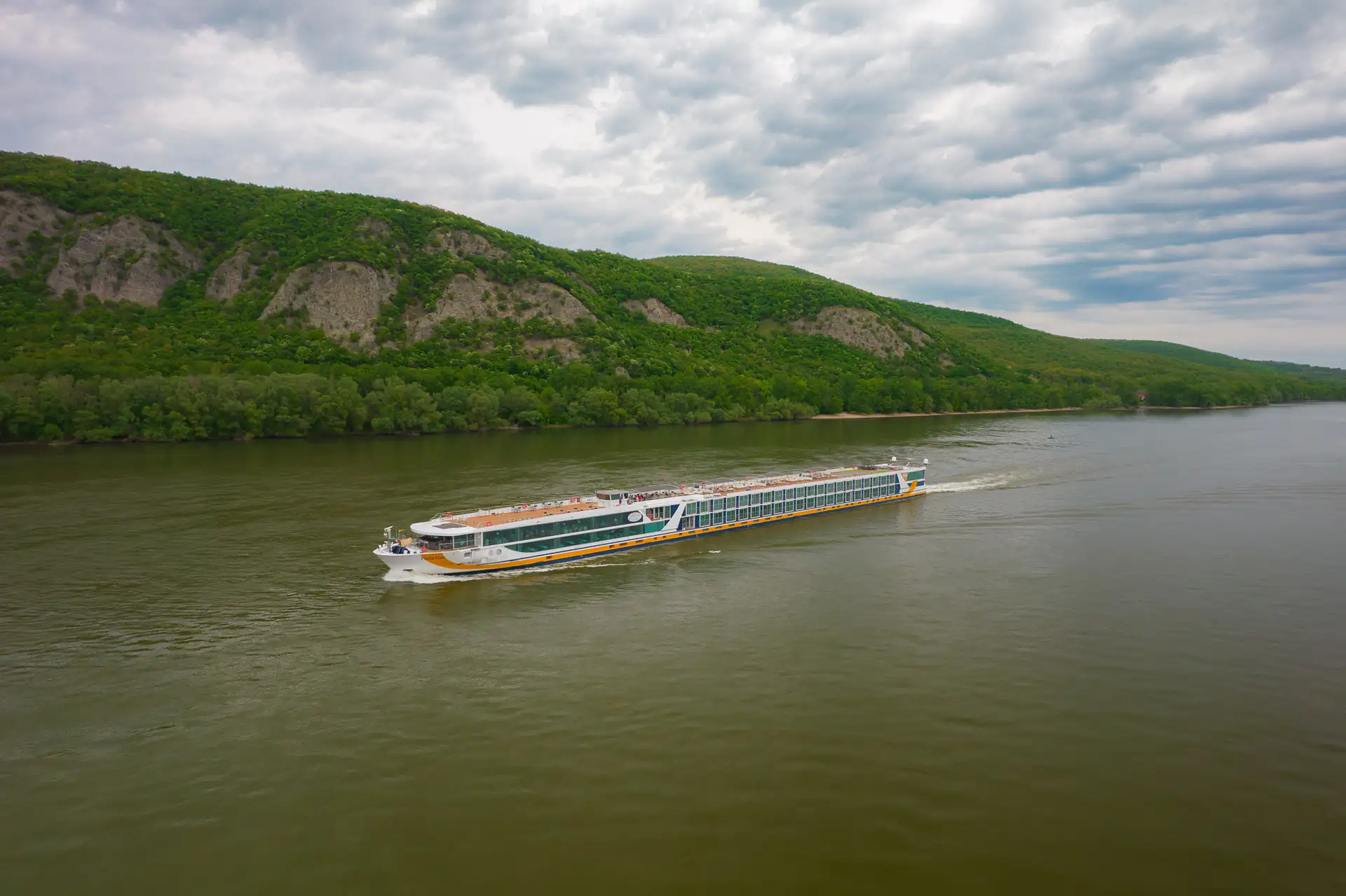 MS VistaSTAR auf ruhiger Donau vor bewaldetem Hügel mit Felsen unter bewölktem Himmel.