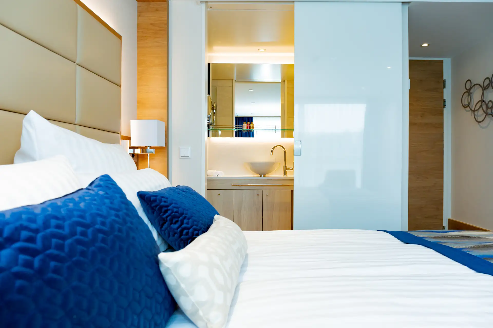 MS VistaSTAR: Bett mit weißen Laken und blauen Kissen vor einem gepolsterten Kopfteil, im Hintergrund ein Badezimmer mit Waschbecken und Spiegel.