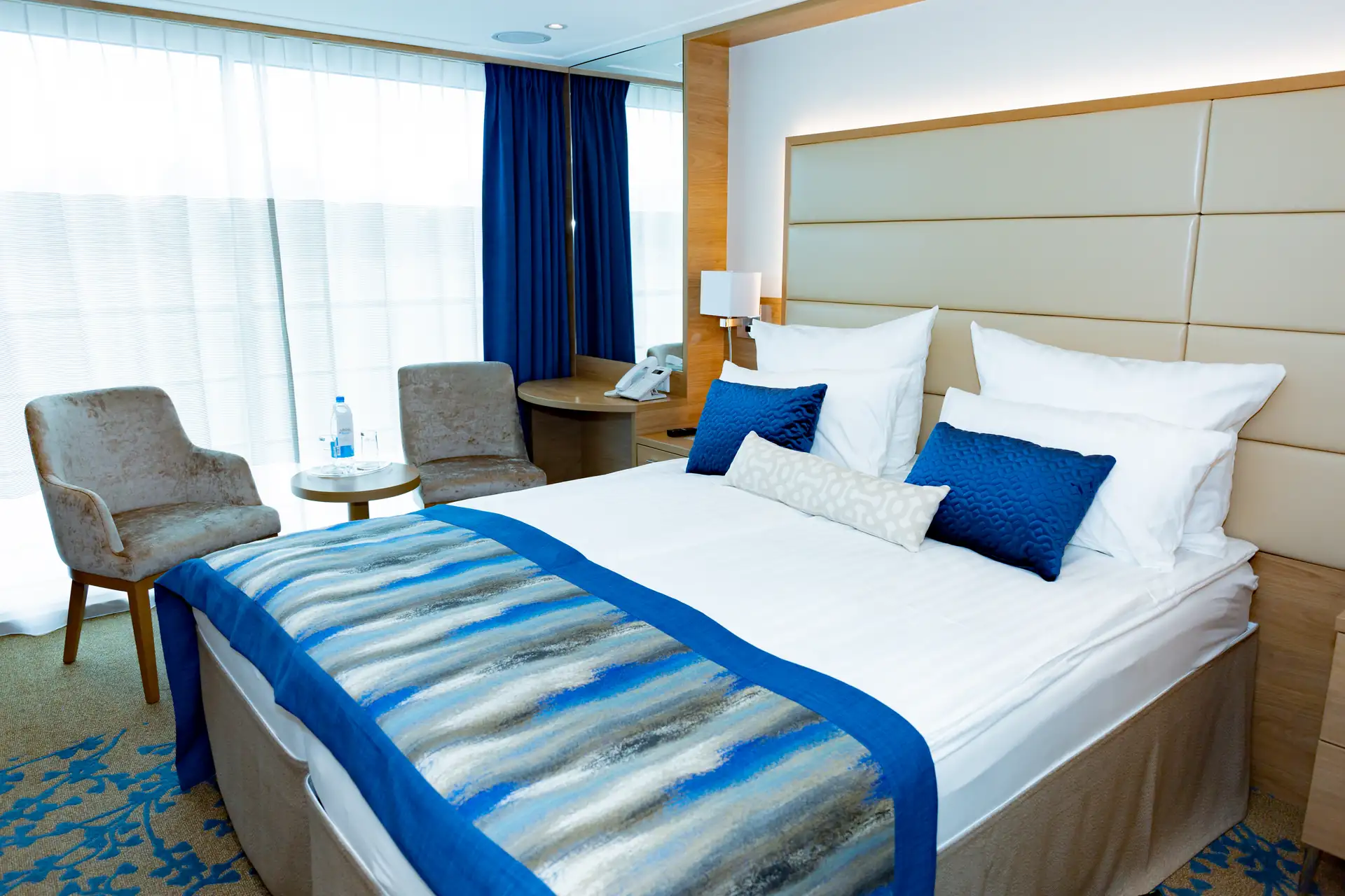 MS VistaSTAR Kabine auf dem Mittel-/Oberdeck mit Doppelbett, blauer Tagesdecke und Kissen, zwei grauen Sesseln, rundem Tisch mit Wasserflasche und Telefon auf Nachttisch.