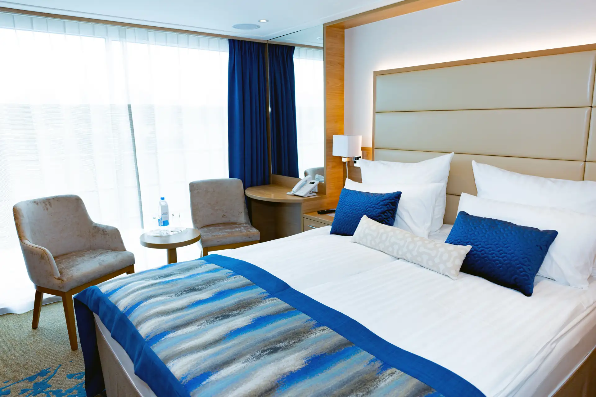 MS VistaSTAR Kabine mit Doppelbett, blauen Kissen und Tagesdecke, zwei grauen Sesseln, rundem Tisch mit Wasserflasche und Telefon auf Holzschreibtisch.