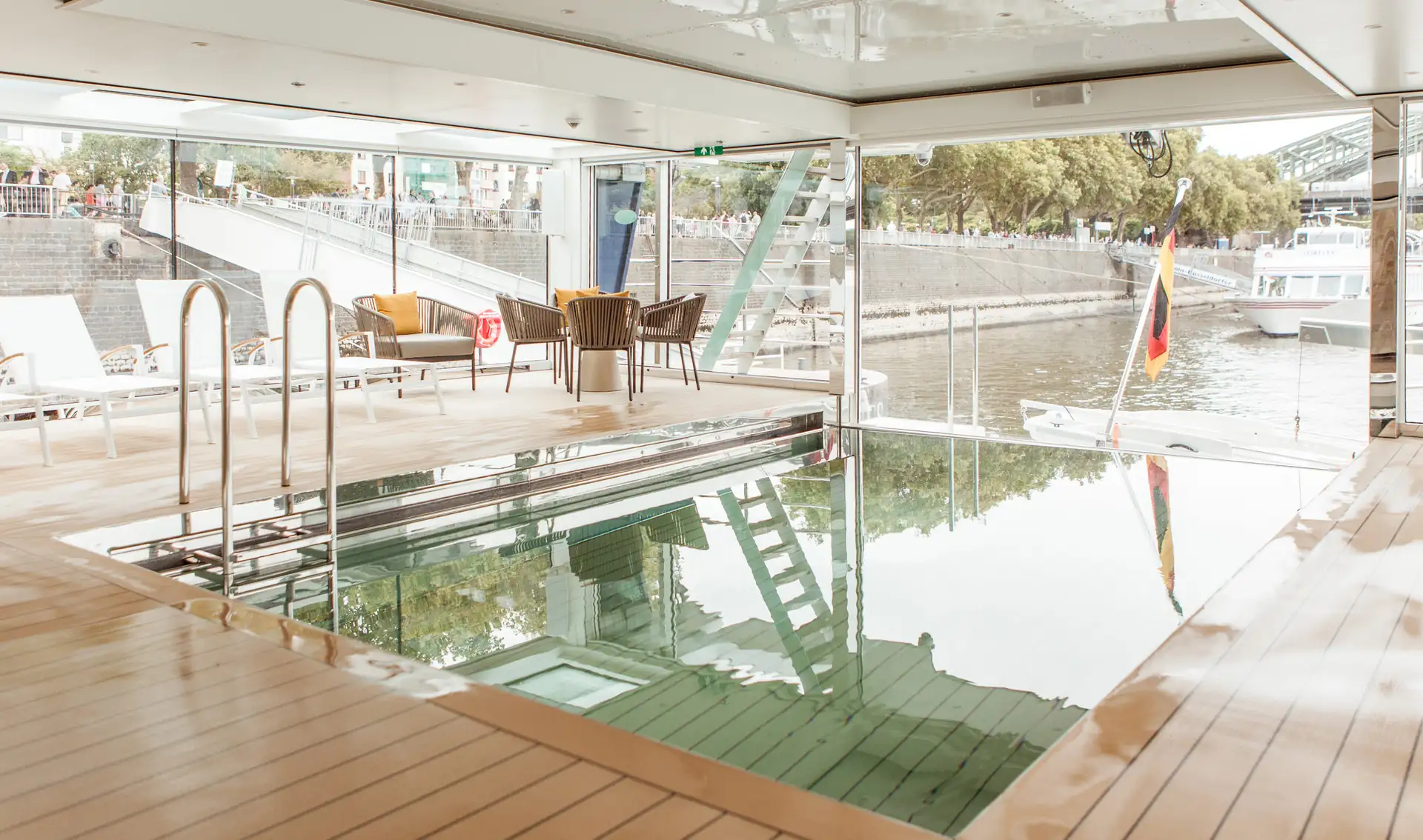 MS VistaSTAR: Schwimmbecken, Holzfußboden, Sitzmöbeln und Blick auf Ufer mit Bäumen und Gebäuden