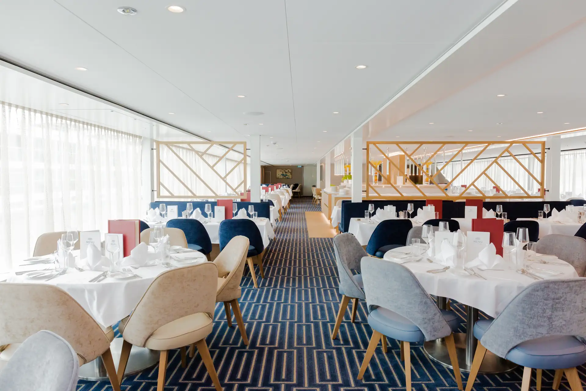 MS VistaSTAR: Heller Speisesaal mit runden Tischen, gedeckt mit weißem Geschirr und Gläsern, gepolsterte Stühle in Beige, Blau und Grau, geometrischer Teppichboden.