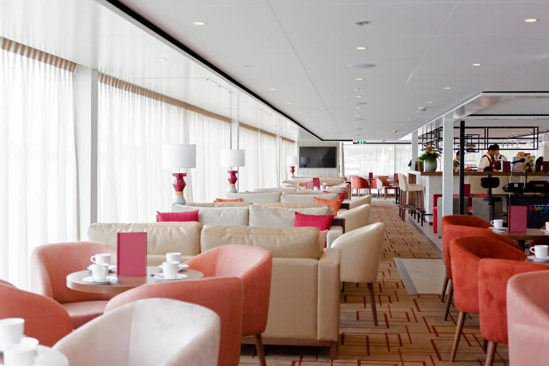 MS VistaSTAR: Heller, moderner Gastraum mit beigen Sofas, roten Sesseln, weißen Tassen auf Tischen und großen Fenstern mit Gardinen.