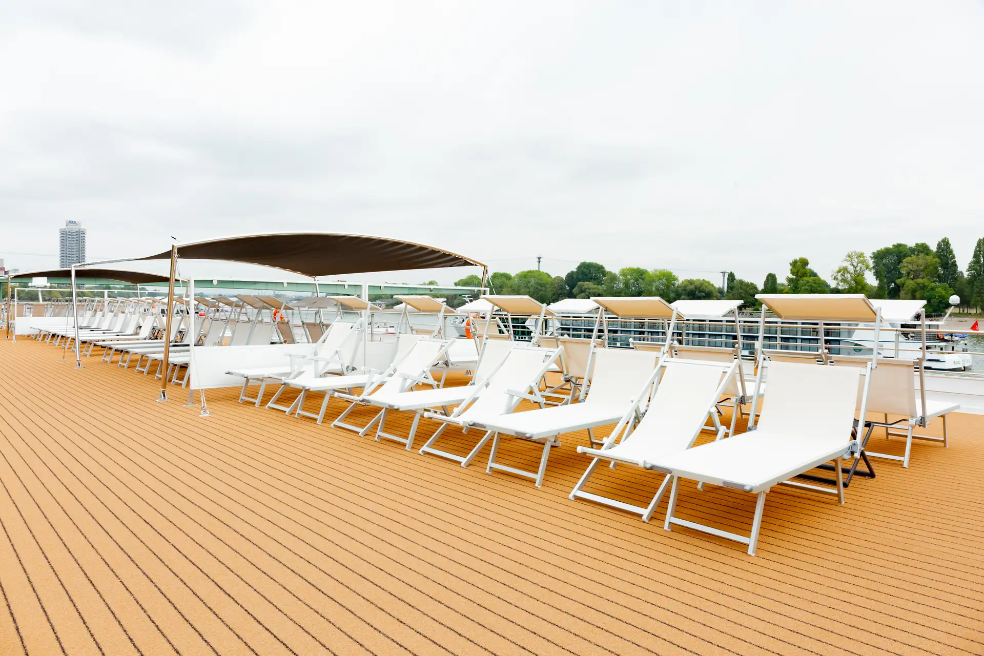 MS VistaSTAR: Reihe weißer Liegestühle auf Holzdeck mit Sonnenschutzdach und Bäumen im Hintergrund.