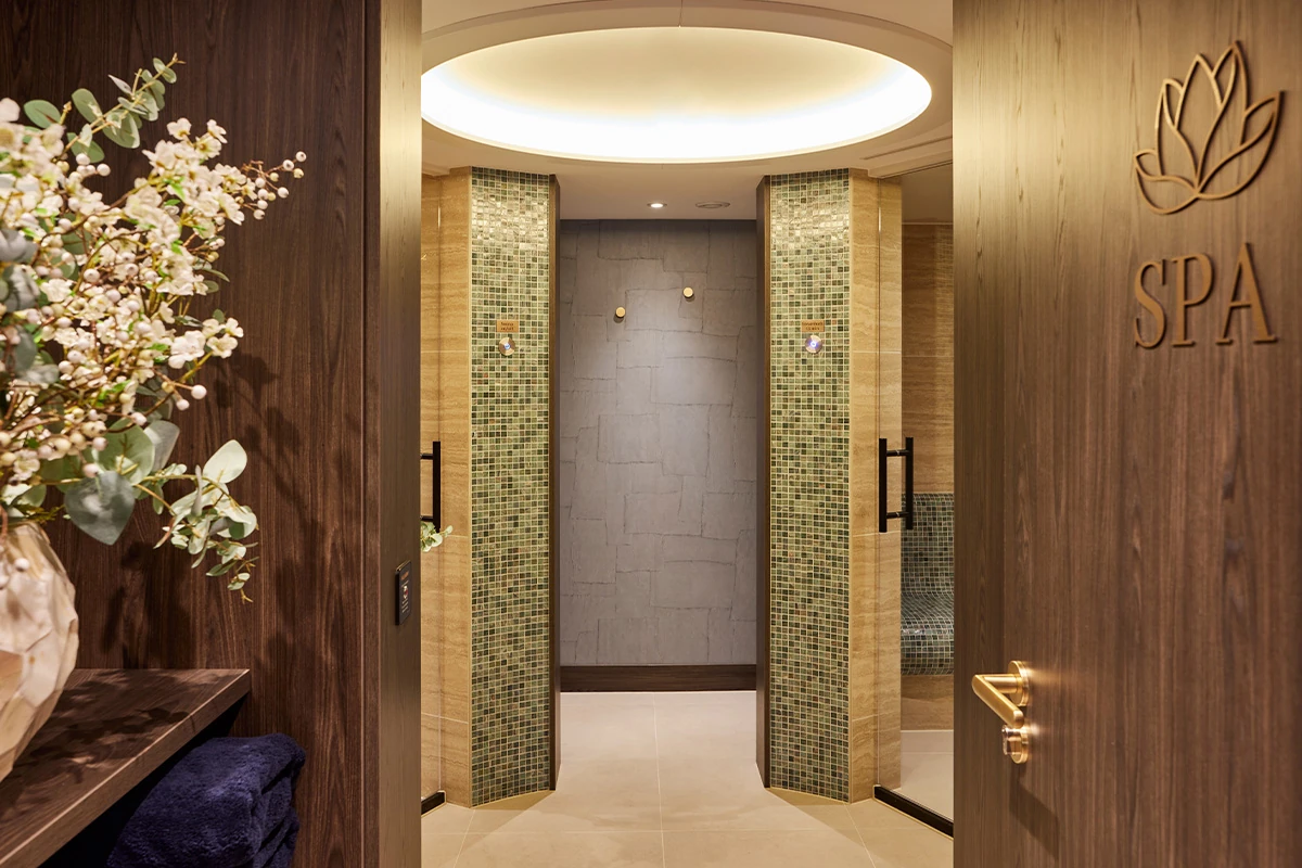 Eingangsbereich eines Spabereichs mit offenem Holztürblatt, das ein goldfarbenes SPA-Logo zeigt, links ein Blumenarrangement auf einem Regal und zwei offene Duschen mit Mosaikfliesen unter einer runden Deckenleuchte