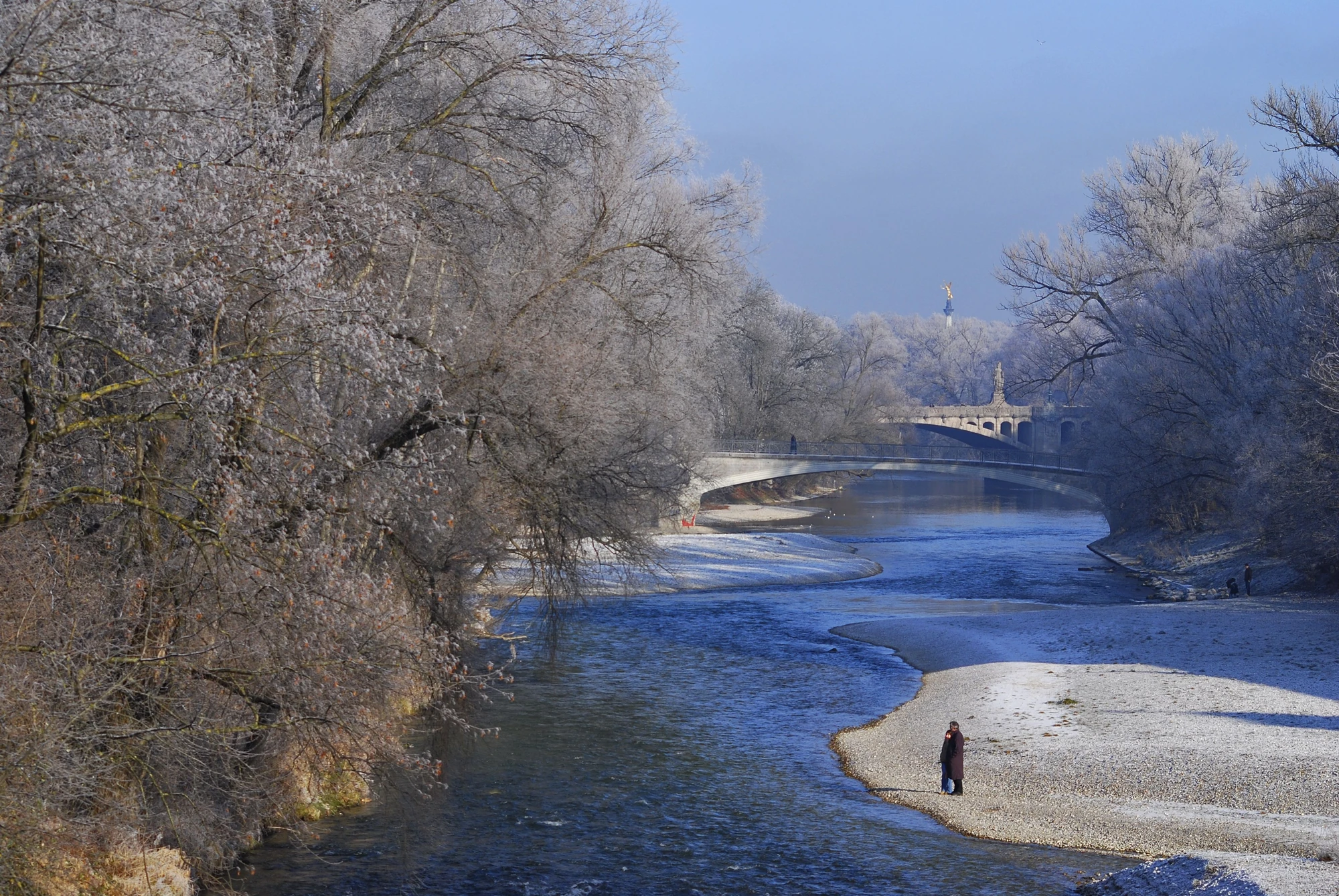 Bick auf die Isarbrücken, ein Pärchen steht am Flußufer und blickt auf die mit Raureif überzogenen Bäume