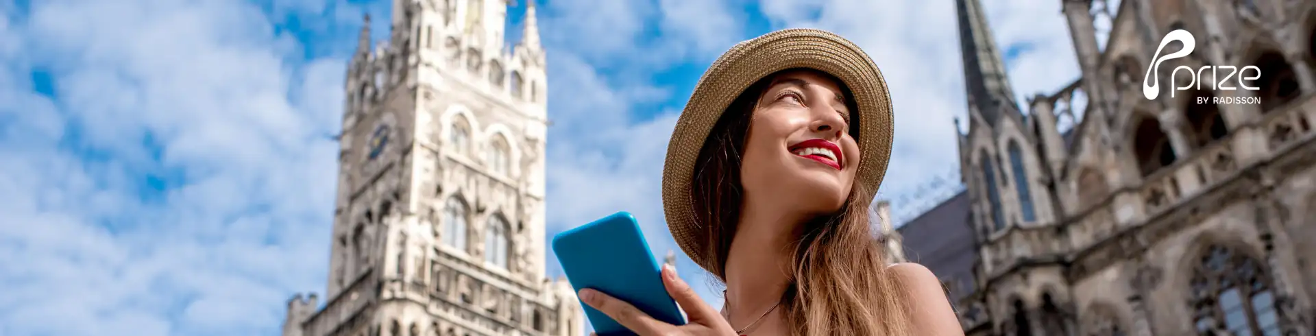 Junge Frau mit Strohhut und Smartphone vor dem neugotischen Turm des Münchner Rathauses bei blauem Himmel
