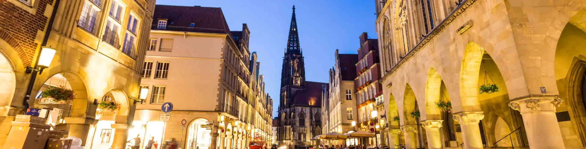 Blick in eine belebte Altstadtstraße Münsters mit historischen Gebäuden und einem hohen Kirchturm im Hintergrund bei Abendlicht