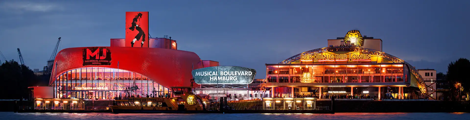 Musicalreisen nach Hamburg im Bannerformat: Stage Theater im Hamburger Hafen am Abend mit den Musicals MJ und Disneys Der König der Löwen.