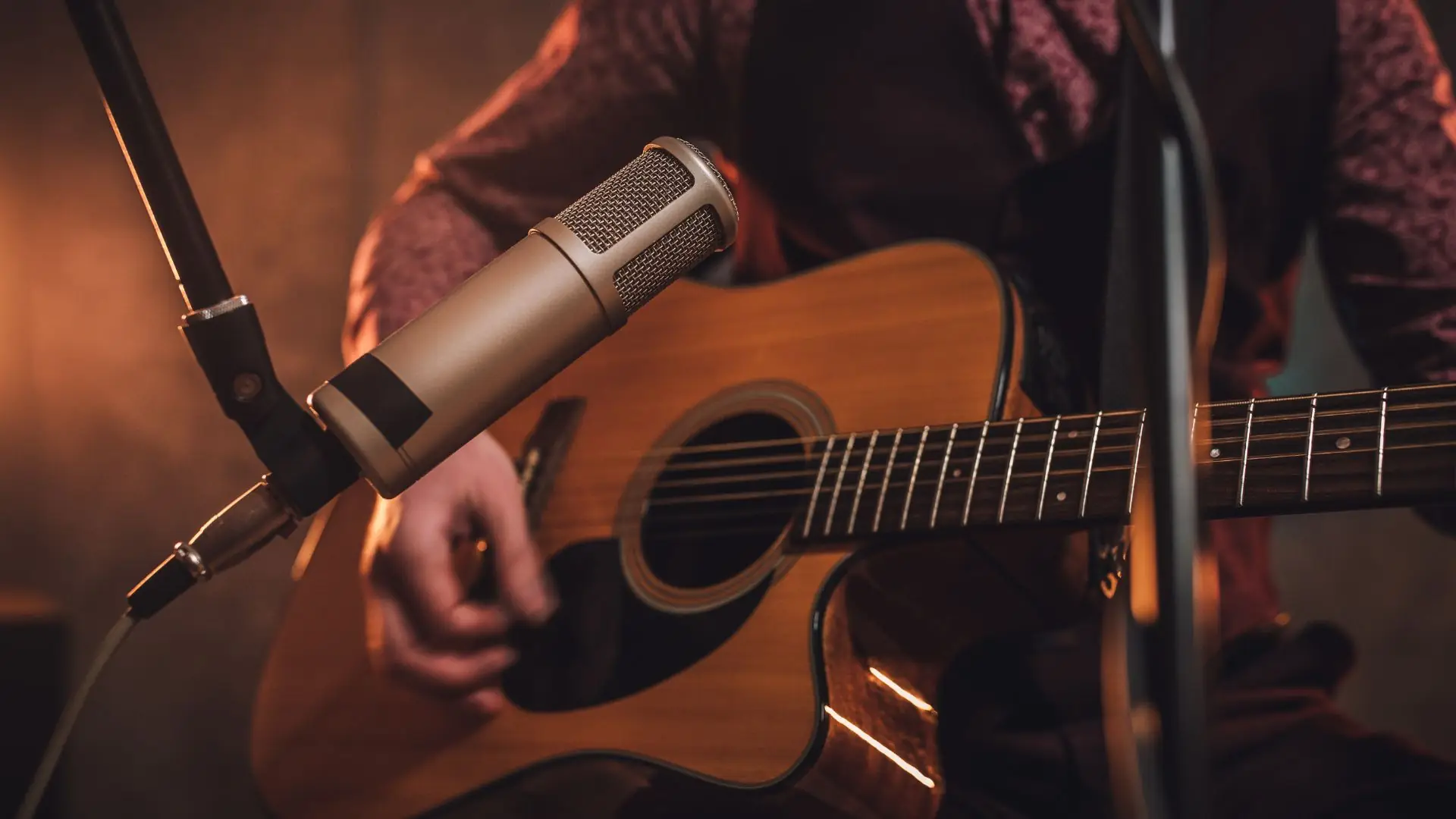 Nahaufnahme einer akustischen Gitarre, die von einer Person mit Hemd und Weste gespielt wird, davor ein Mikrofon an einem Stativ.