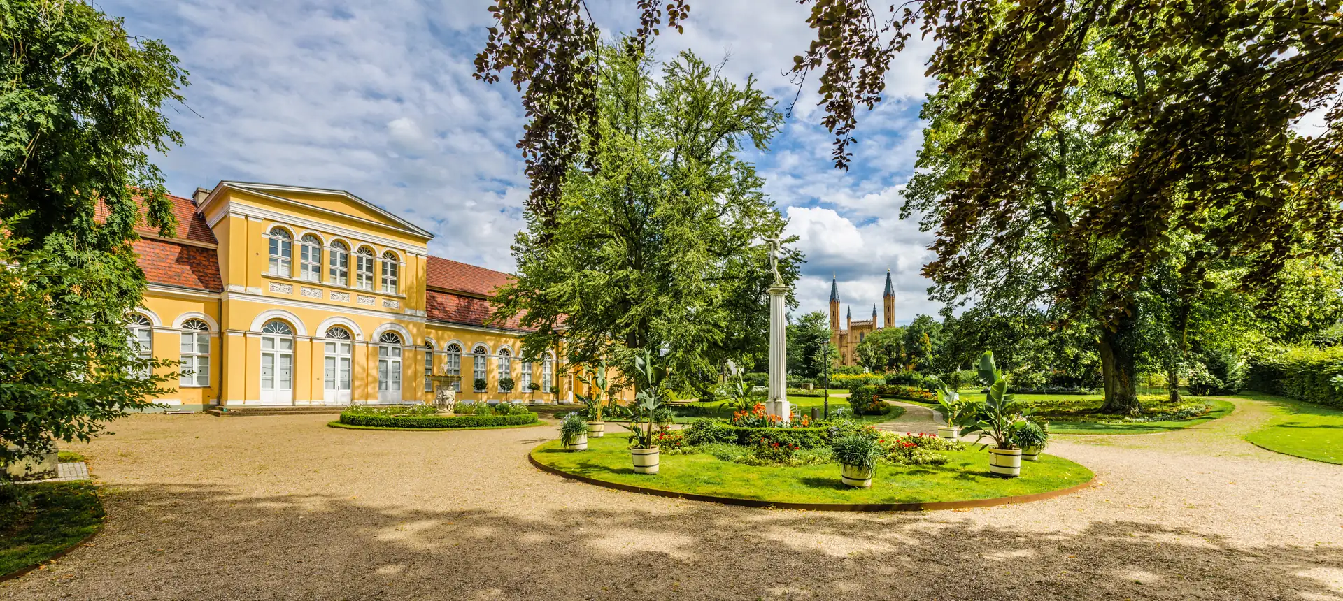 Gelber klassizistischer Pavillon im Schlossgarten Neustrelitz mit umlaufendem Kiesweg, grünen Bäumen und einer runden Blumenrabatte mit weißem Obelisken im Zentrum