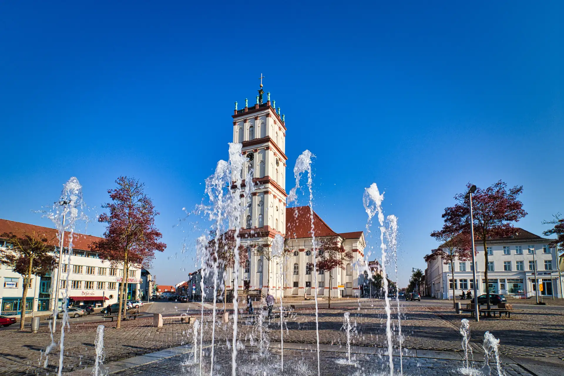 Die Stadtkirche Neustrelitz mit hohem Turm steht auf einem Platz mit mehreren Springbrunnen im Vordergrund und umgeben von Gebäuden und Bäumen mit rotem Laub