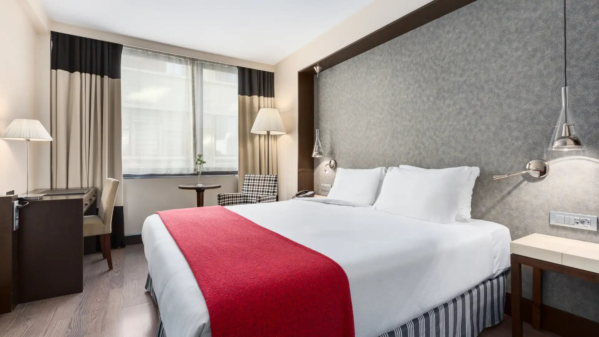 NH Brüssel City Centre Hotelzimmer mit großem Doppelbett, weißer Bettwäsche und rotem Überwurf, zwei Nachttischen mit Lampen, Schreibtisch und Fenster mit Vorhängen