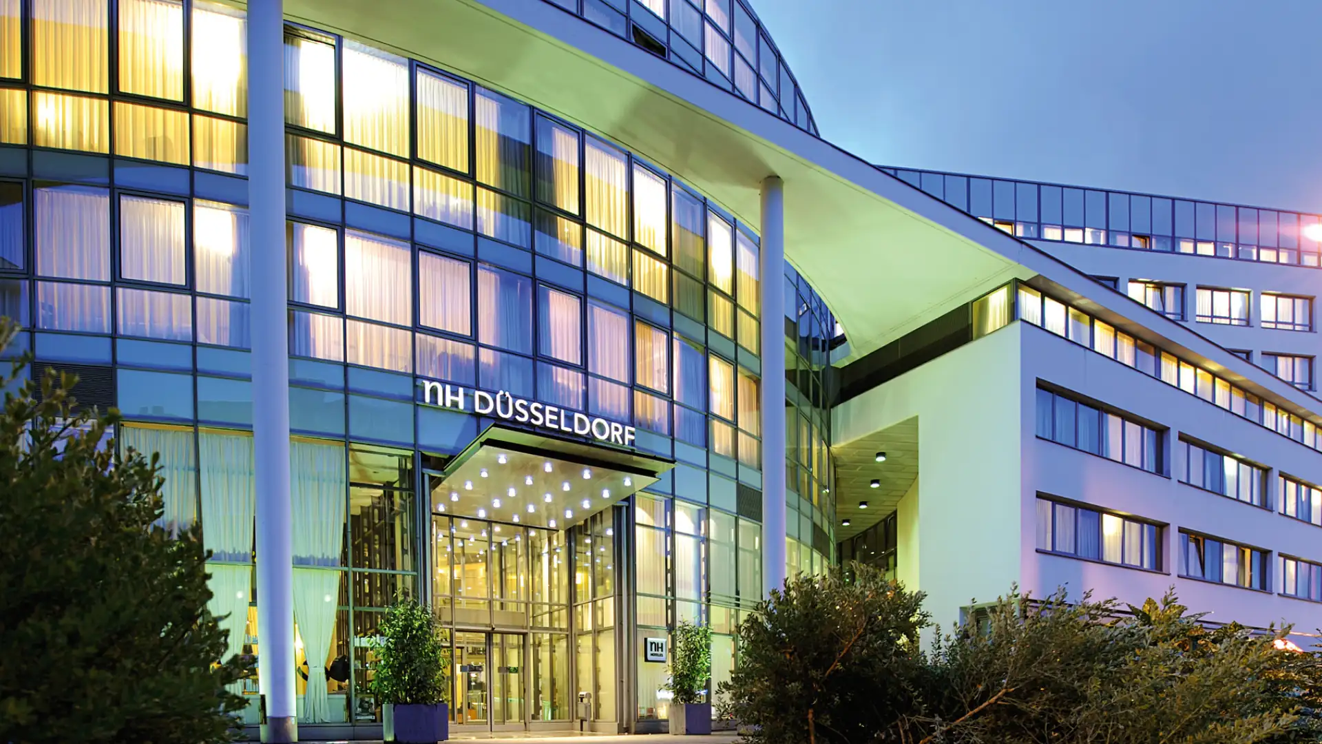 NH Düsseldorf im Kachelformat: Eingang des NH Hotels in Düsseldorf mit beleuchtetem Schriftzug und moderner Glasfassade.