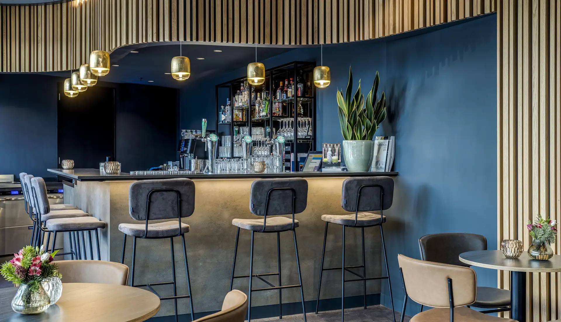 Bar mit dunklem Tresen, vier hohen Barhockern, blauen Wänden, mehreren goldfarbenen Hängeleuchten und Pflanzen auf dem Tresen und Tischen.