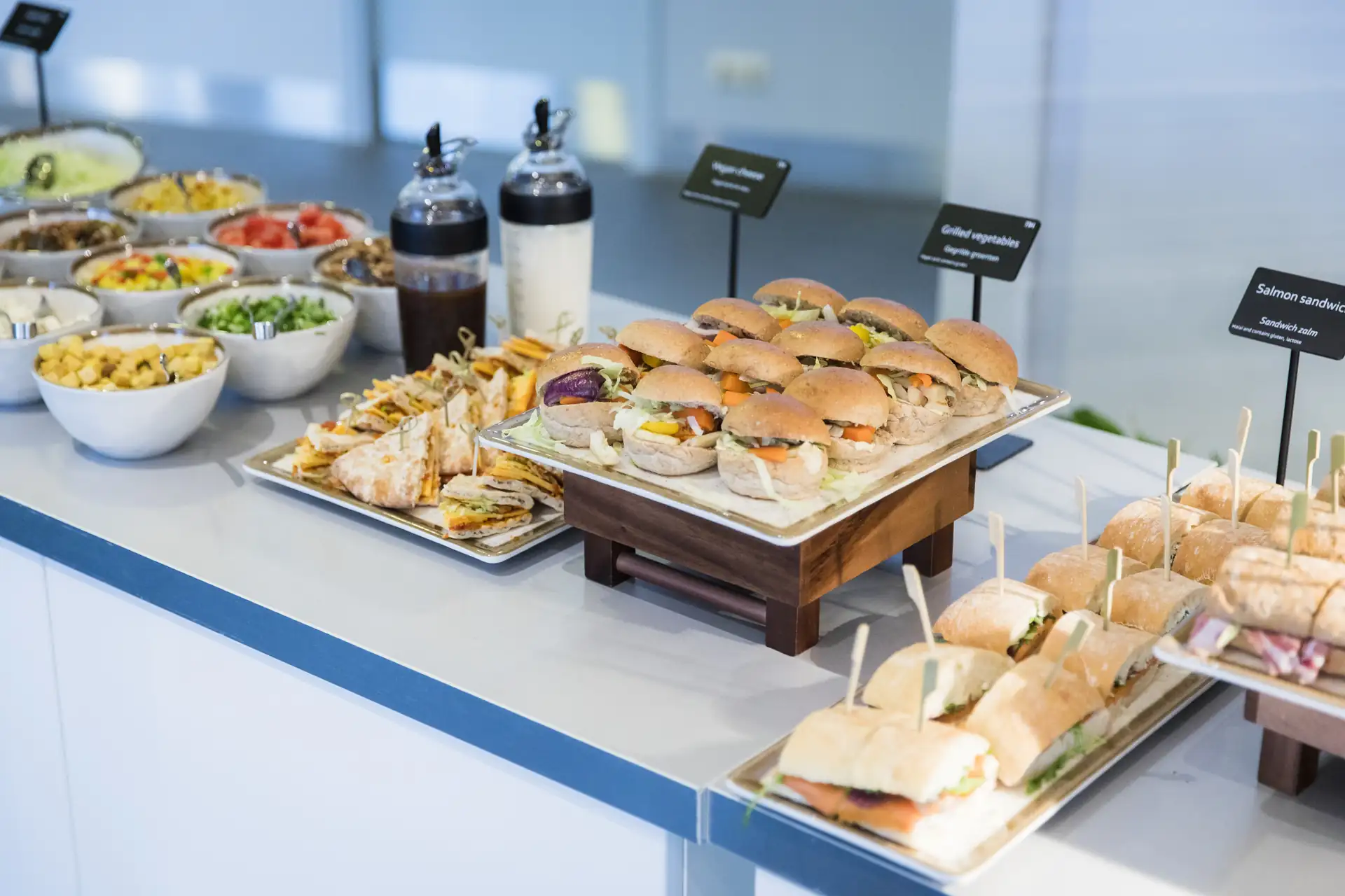 Verschiedene belegte Brötchen und Sandwiches auf Tabletts, daneben Schalen mit Salat und Dressings in Flaschen.