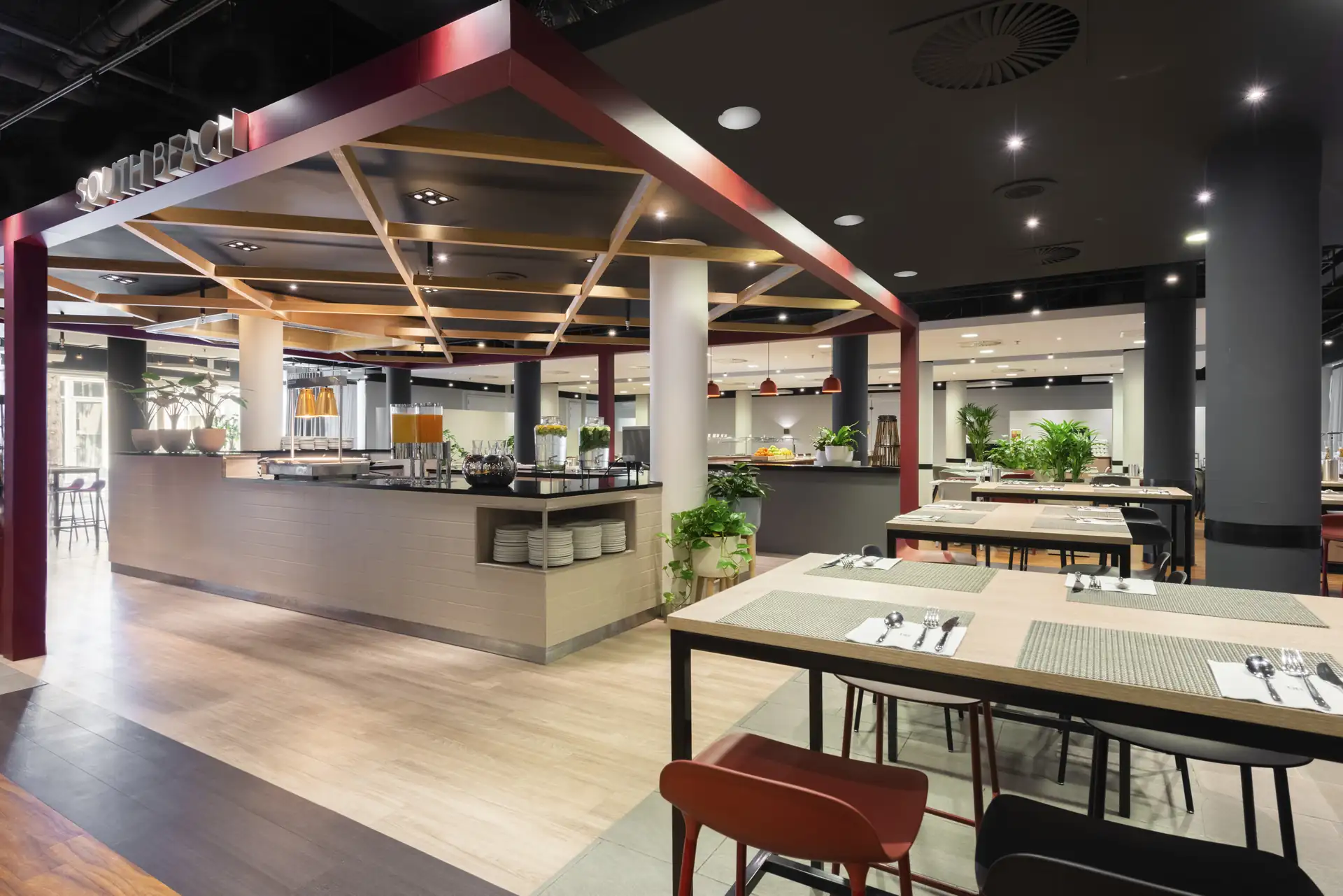 Modern eingerichteter Restaurantbereich mit Tischen, Stühlen, Pflanzen und offenem Buffetbereich unter einer Decke mit sichtbaren Holzbalken.