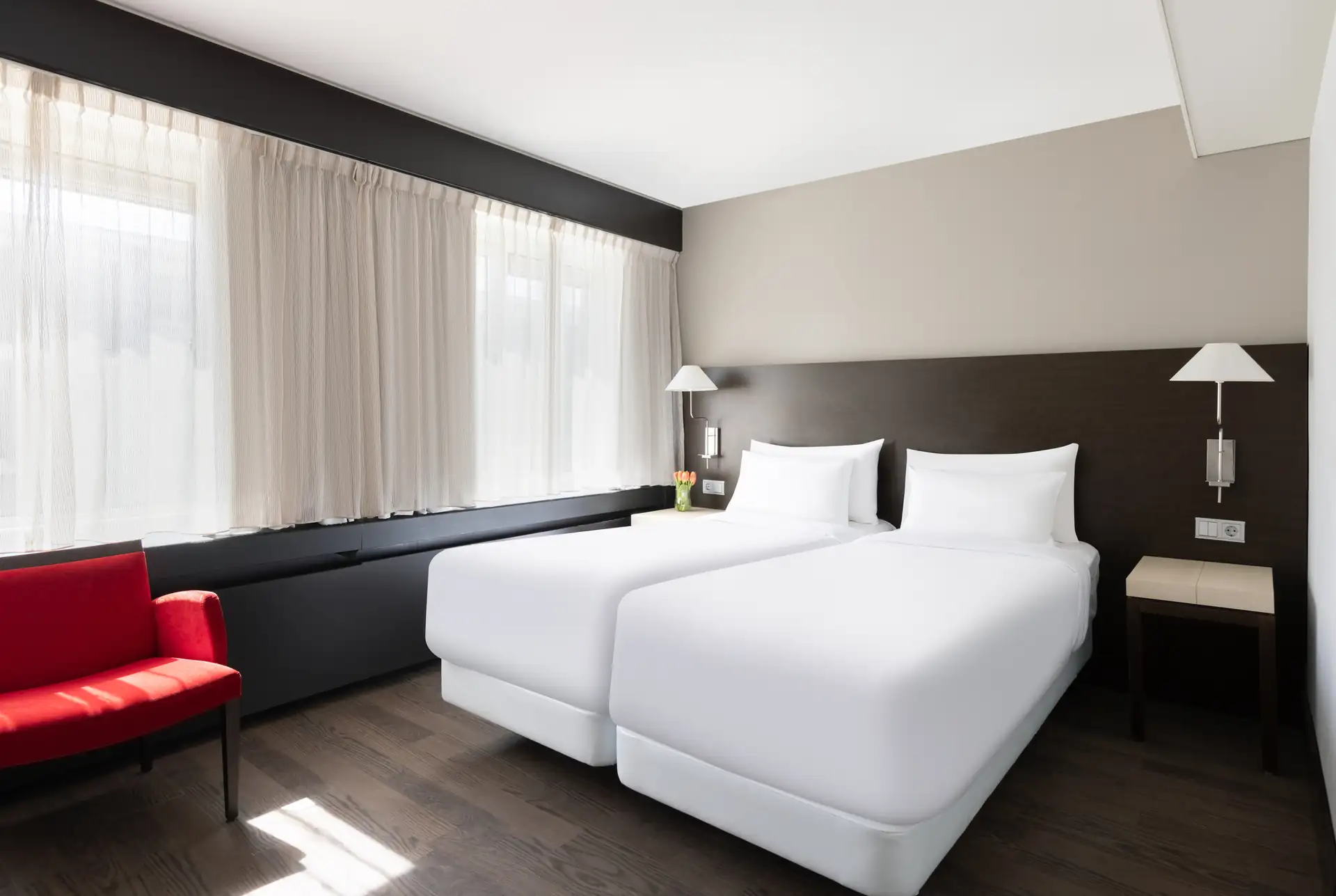 Superior Doppelzimmer mit zwei Einzelbetten, weißen Bettbezügen, Nachttischlampen und rotem Sessel am Fenster mit weißen Vorhängen.