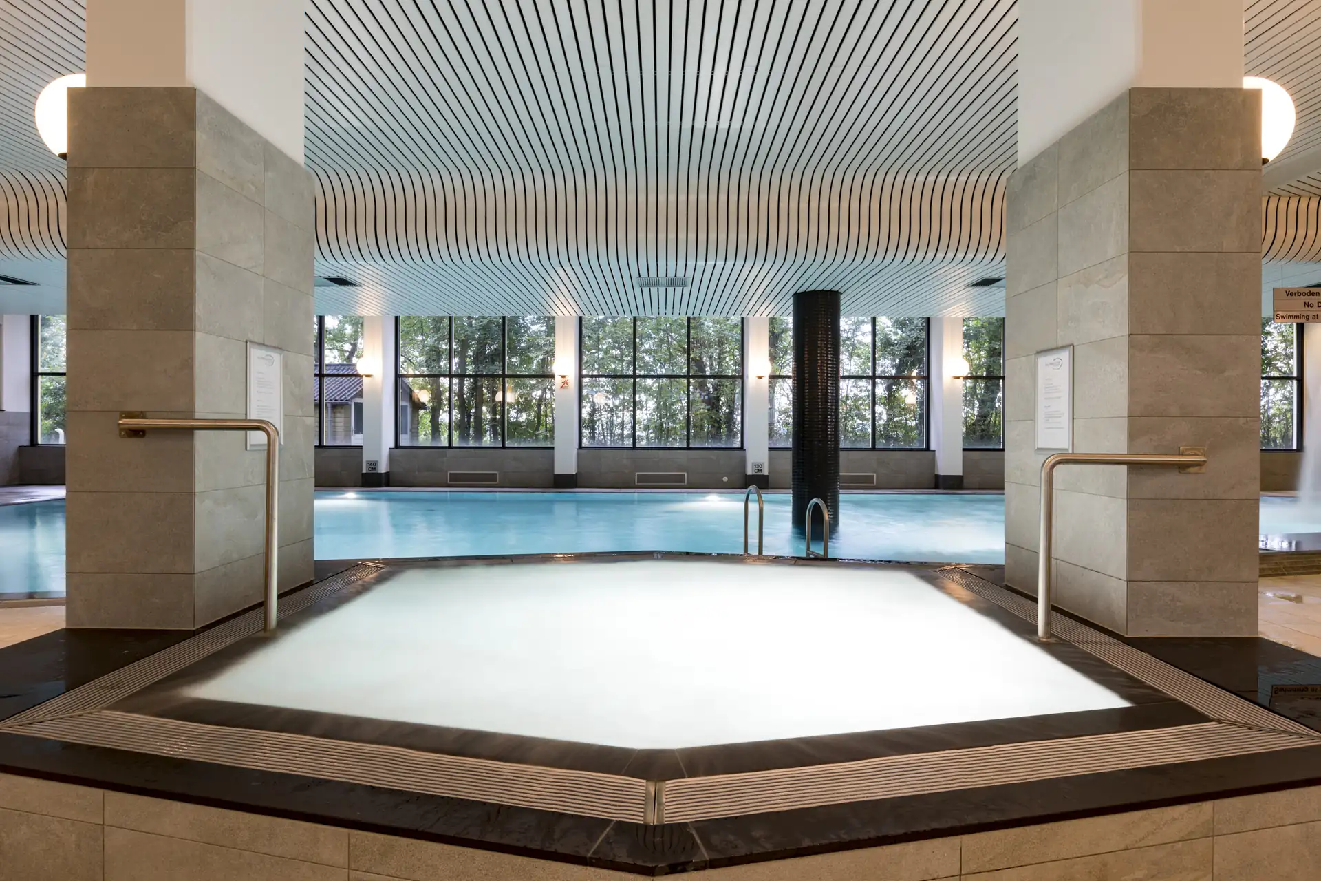 Sechseckiger Whirlpool in einem Schwimmbad, große Fenster und wellenförmige Decke sichtbar.