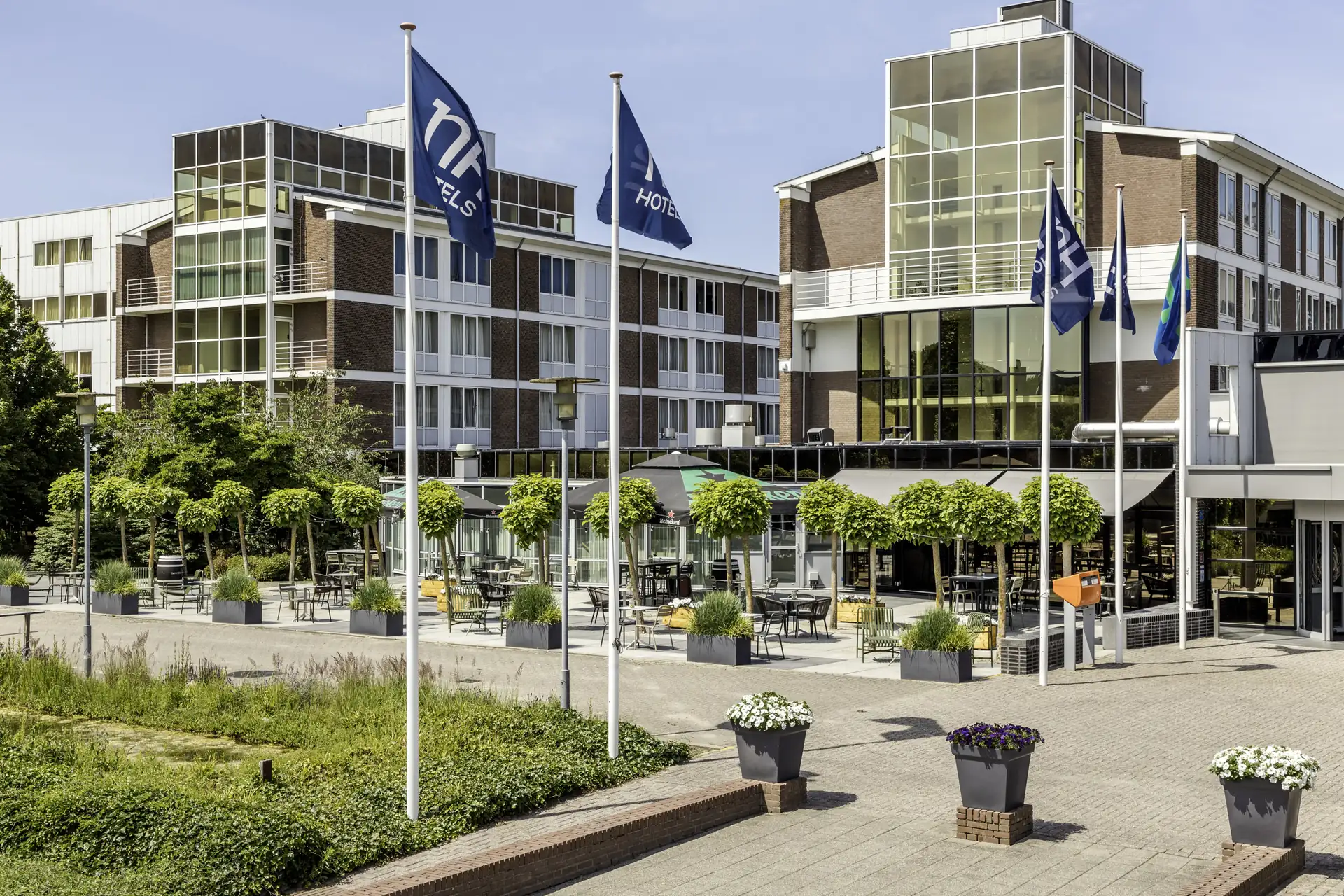 Mehrstöckiges Hotelgebäude mit mehreren blauen Fahnen, die das Wort 'nh HOTELS' zeigen, umgeben von Bäumen und bepflanzten Töpfen.
