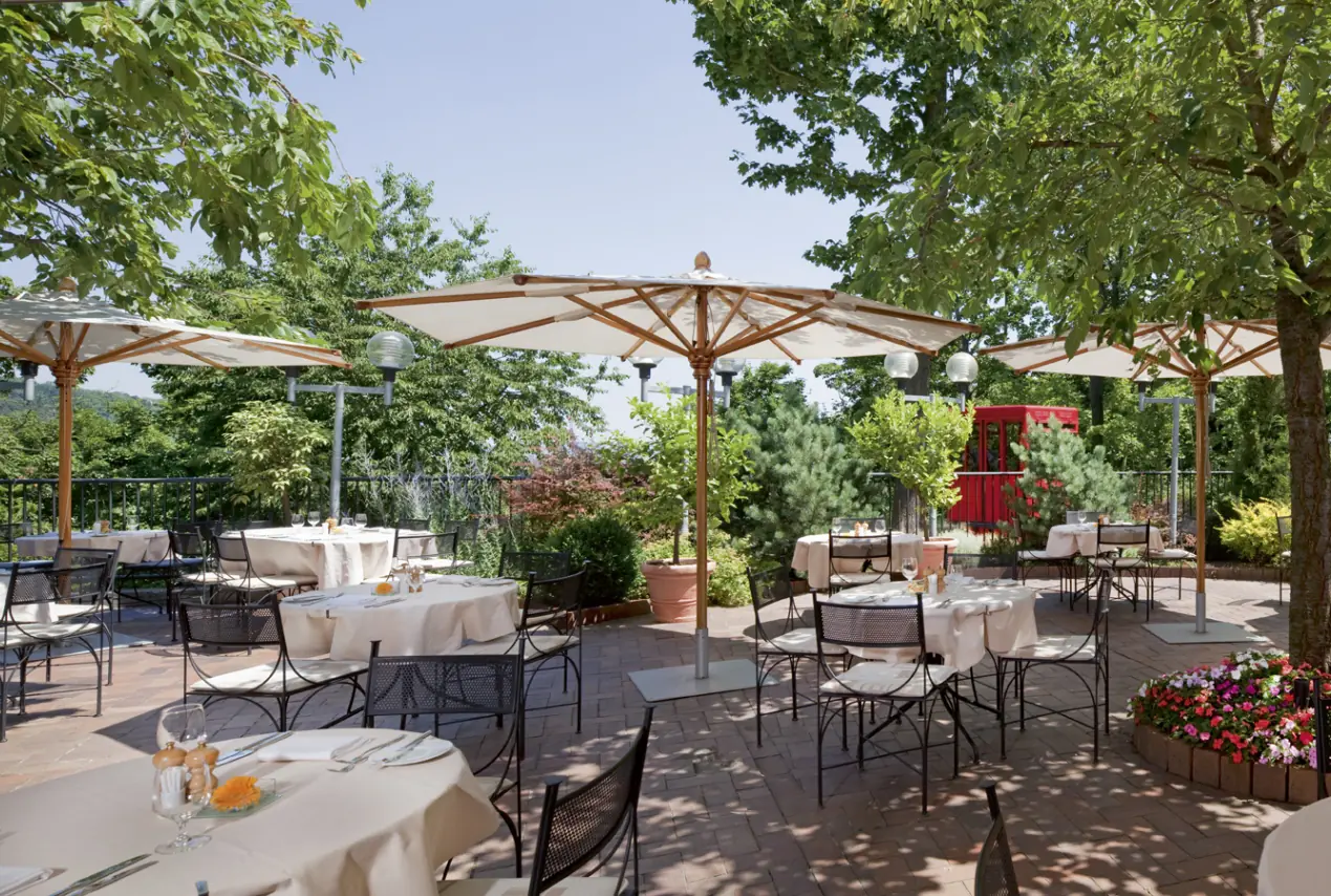 Terrasse des Restaurants Il Giardino mit Sonnenschirmen und gedeckten Tischen vom Schwesternhotel NH Collection in Prag. Im Hintergrund ist die rote Kabine der Seilbahn zu sehen.
