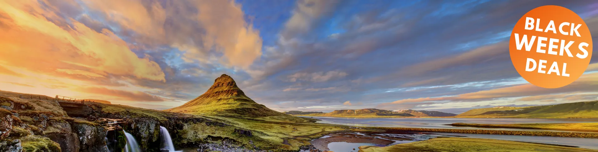 Ein Panoramablick auf den grasbewachsenen Gipfel des Berges Kirkjufell neben einem kleinen Wasserfall und einem Fluss bei Sonnenuntergang, mit einem farbenfrohen Himmel und vereinzelten Wolken.