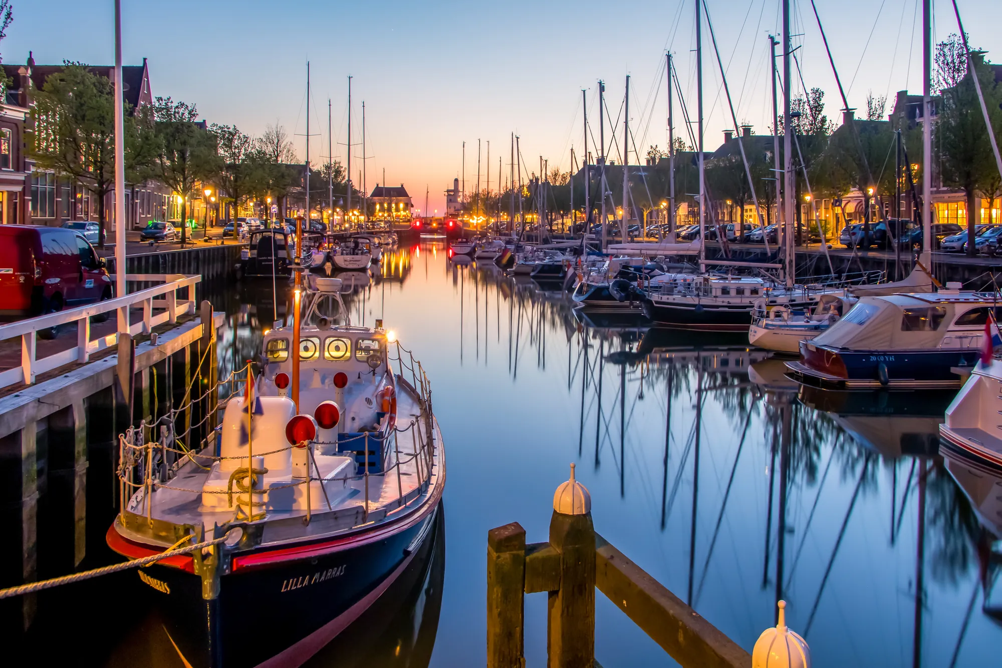 Hafen von Harlingen am Abend, wo angelegte Boote sanft im Wasser schaukeln. Die Stadtbeleuchtung spiegelt sich in der ruhigen Wasseroberfläche und verleiht der Szenerie einen bezaubernden Charme.